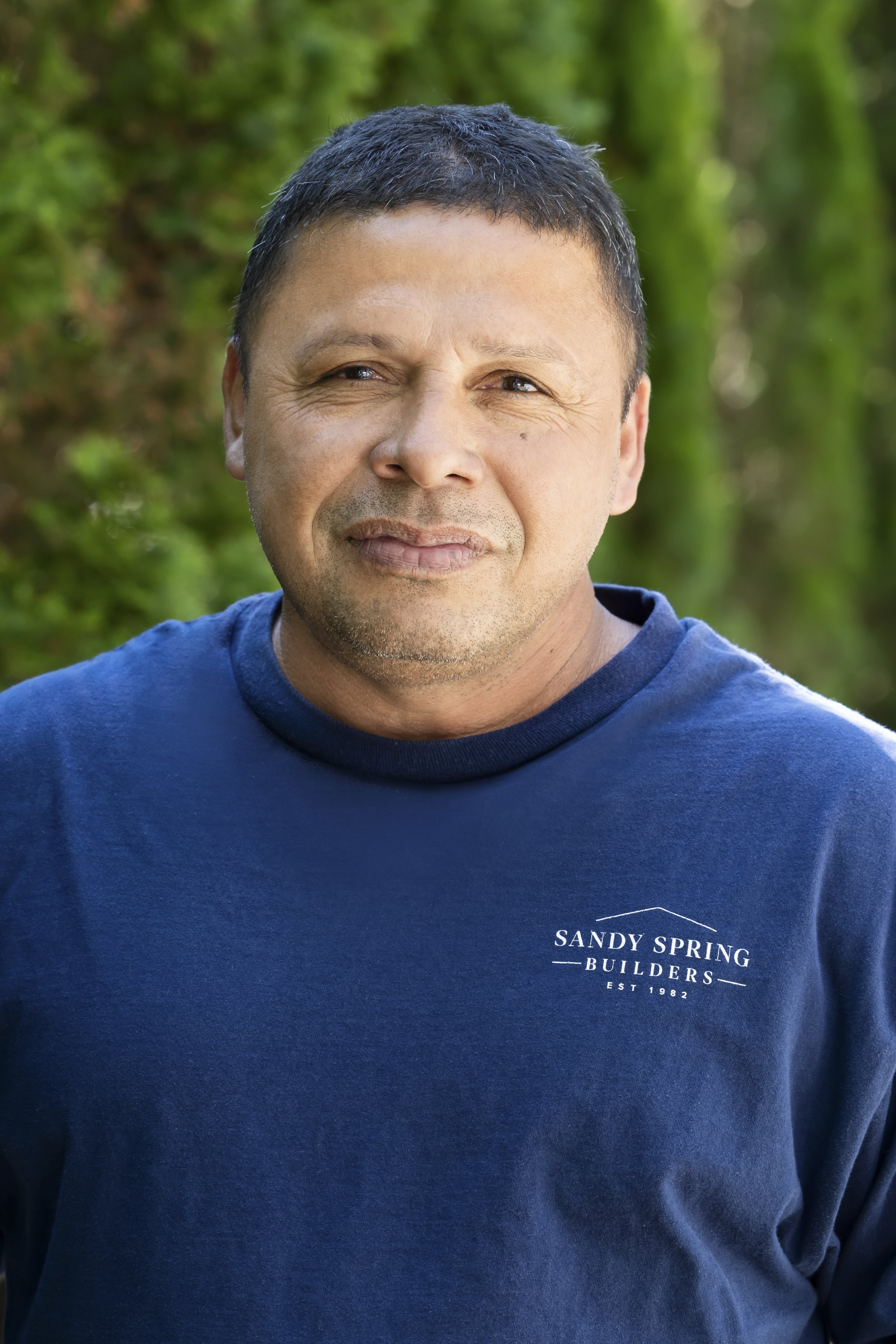 A man with short dark hair and a slight smile, wearing a blue T-shirt with the text 'SANDY SPRINGS BUILDERS EST 1982' on it, outdoors with a blurred green background of trees and plants.