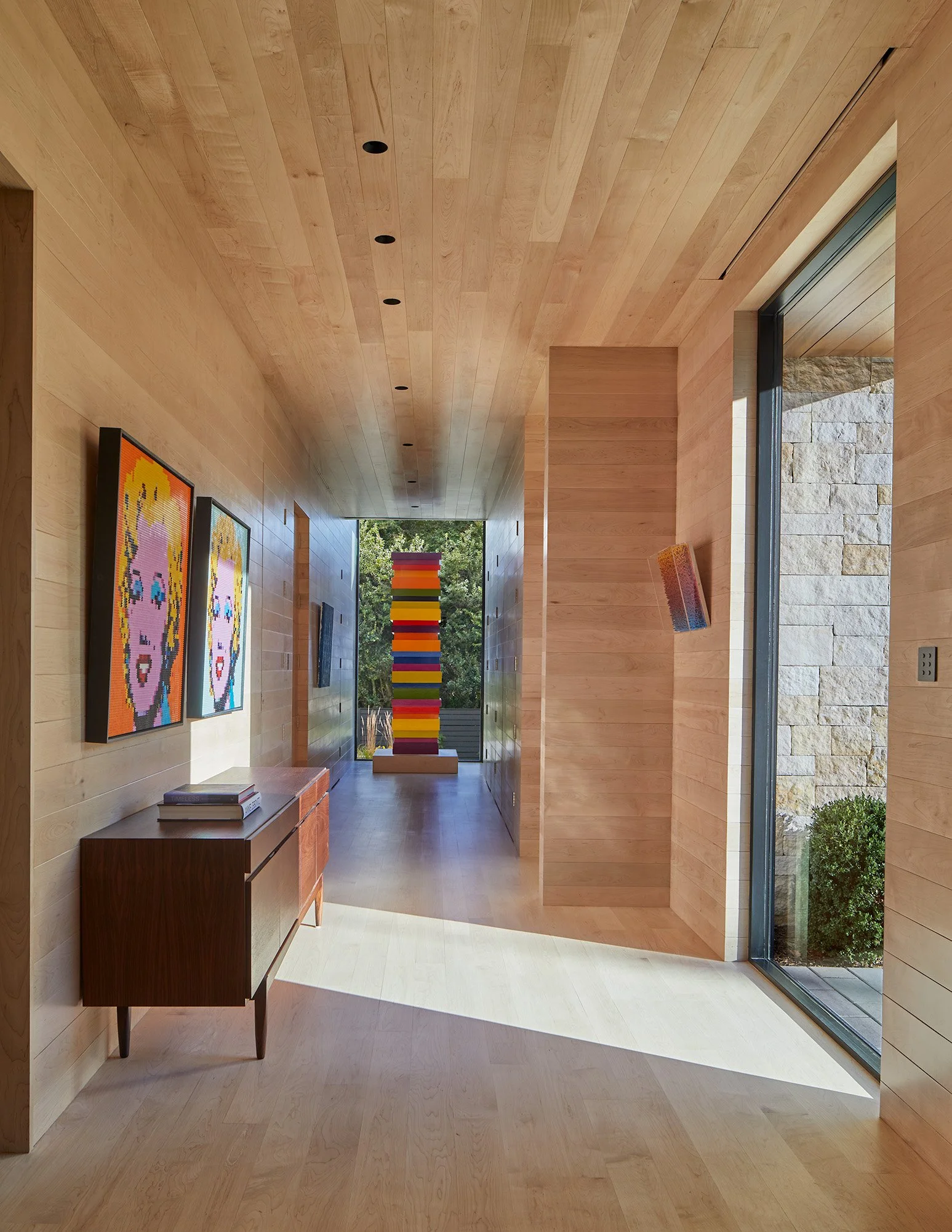 A modern interior corridor with wooden walls, ceiling, and flooring, featuring artwork on the walls and a large window showing greenery outside.