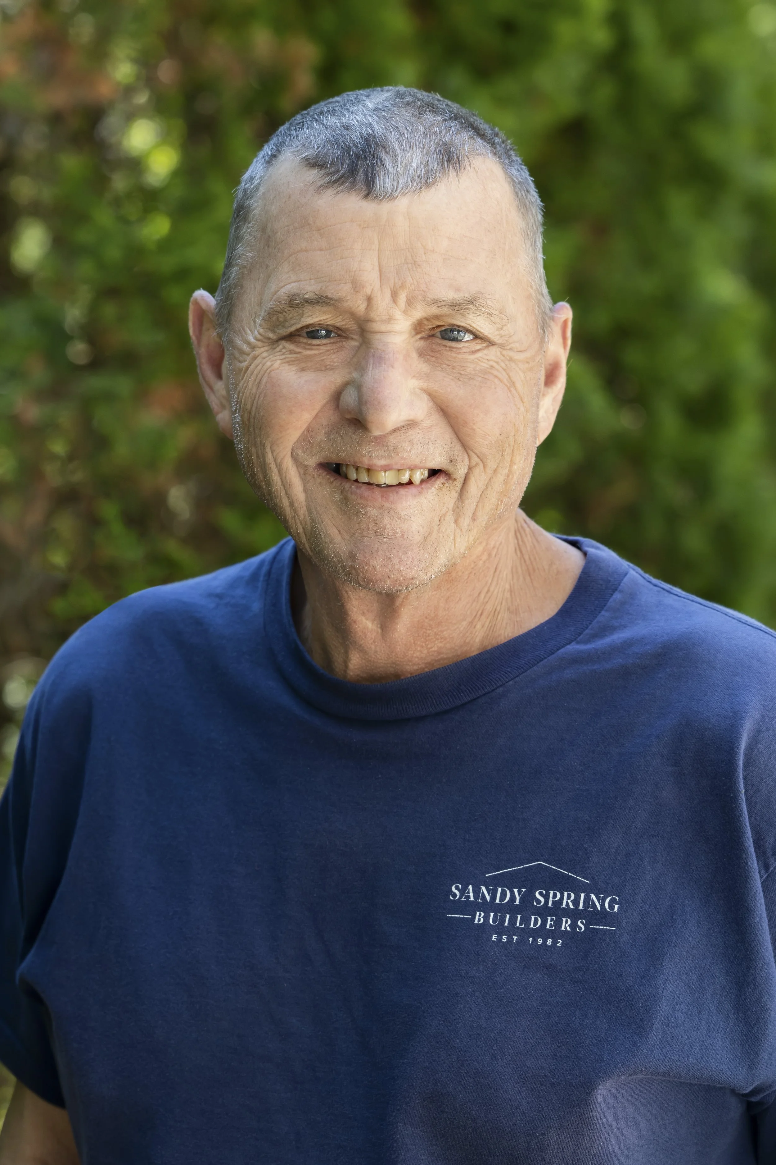 A smiling middle-aged man with short gray hair, wearing a dark blue T-shirt that says 'SANDY SPRING BUILDERS' on it, standing outdoors with a blurred green background of trees and foliage.