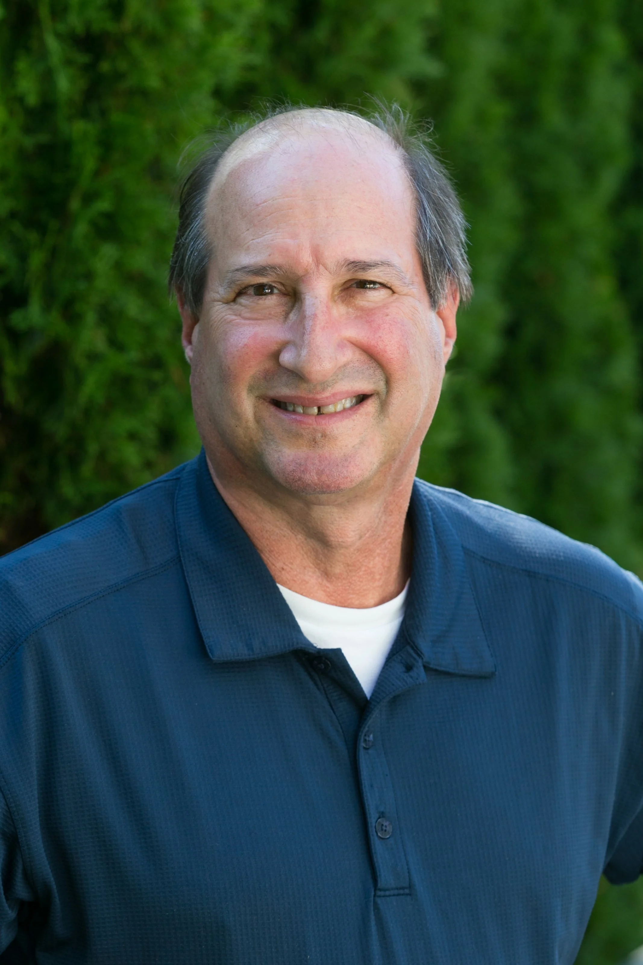 Close-up of smiling middle-aged man with short hair and blue polo shirt, standing outdoors in front of green foliage.