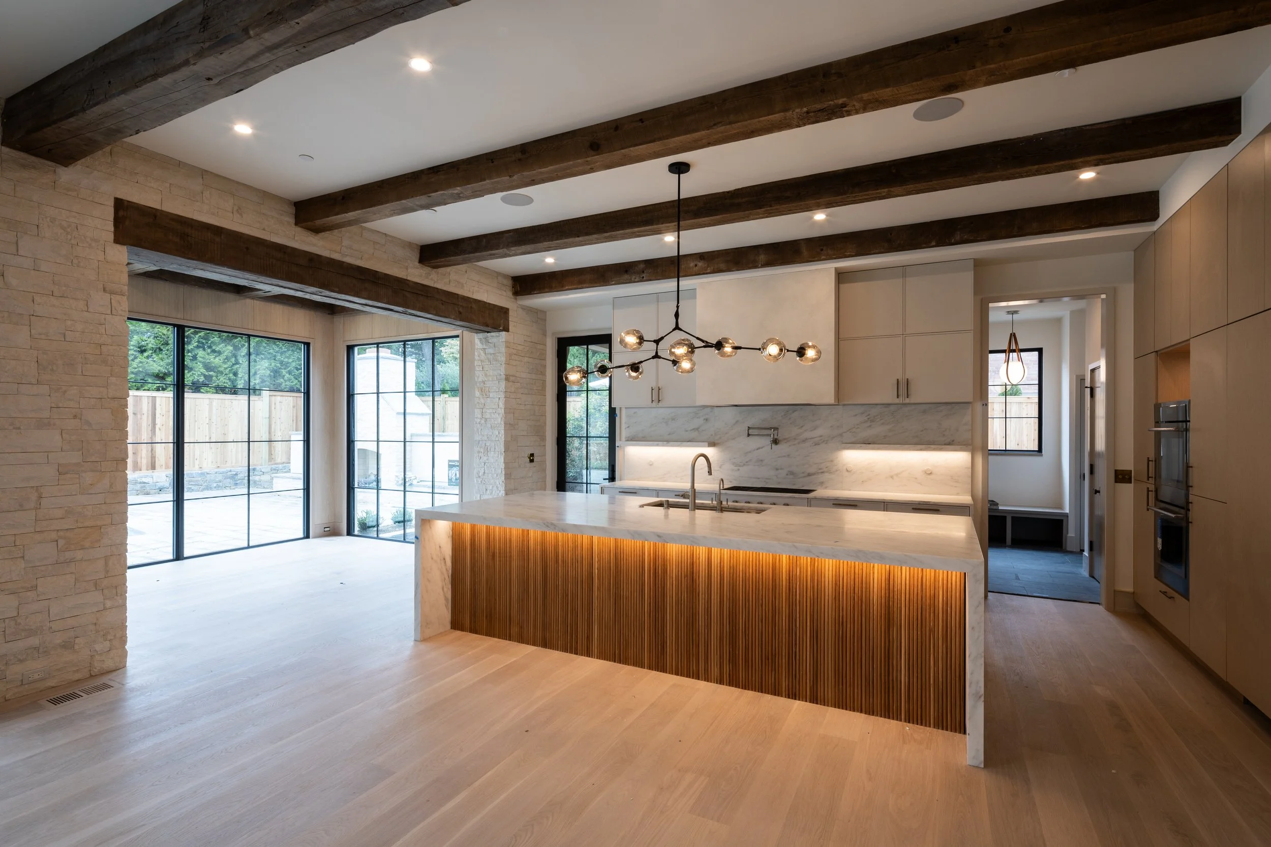 Modern kitchen with beige cabinets, marble countertops, and a kitchen island with wooden paneling, illuminated by contemporary pendant lights with glass globes. Large sliding glass doors open to an outdoor space with a wooden fence.