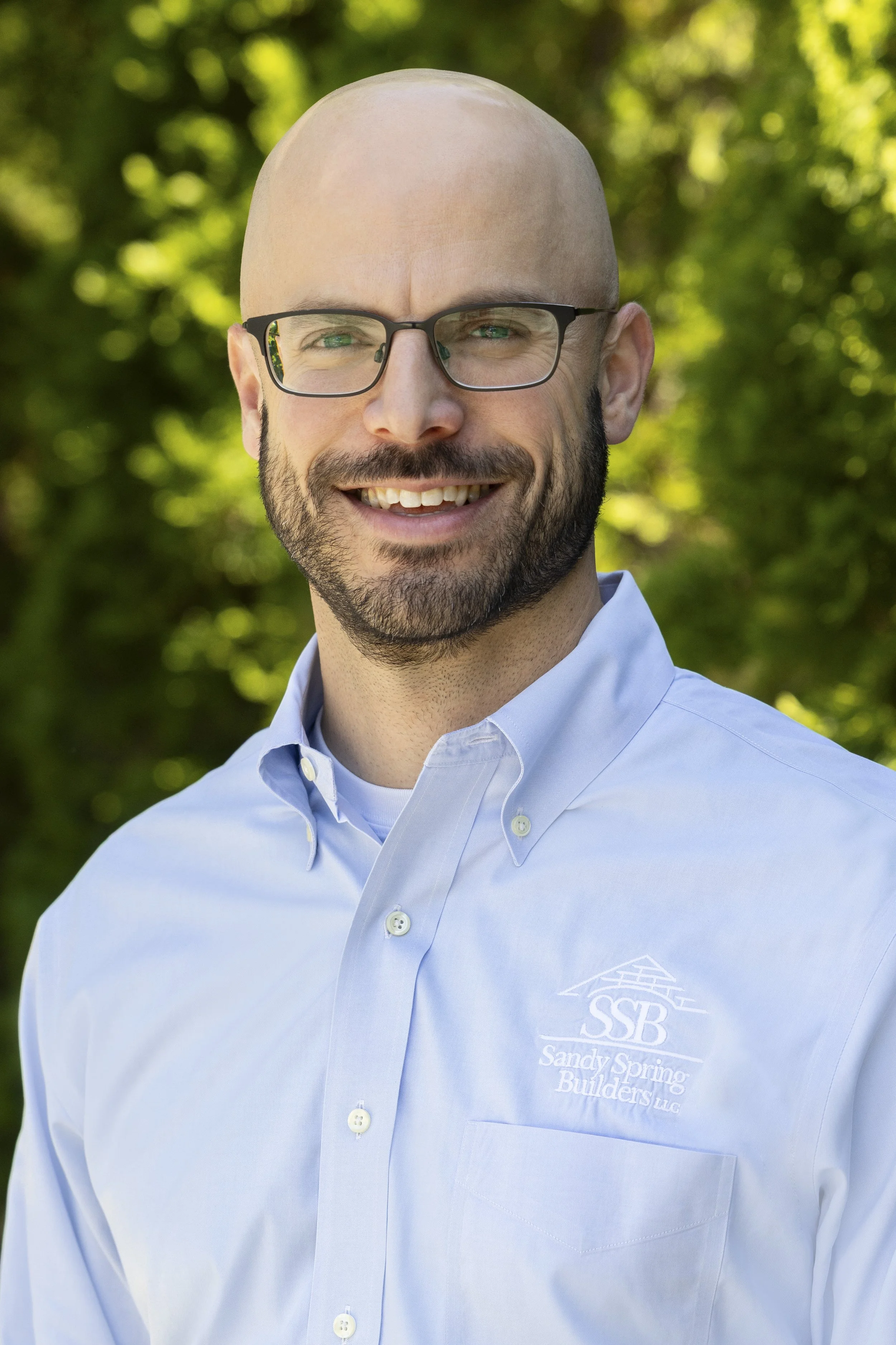 Man with glasses, beard, and bald head wearing a light blue collared shirt with a logo that reads "SSB Sandy Spring Builders LLC" on the chest, outdoors with green foliage in the background.