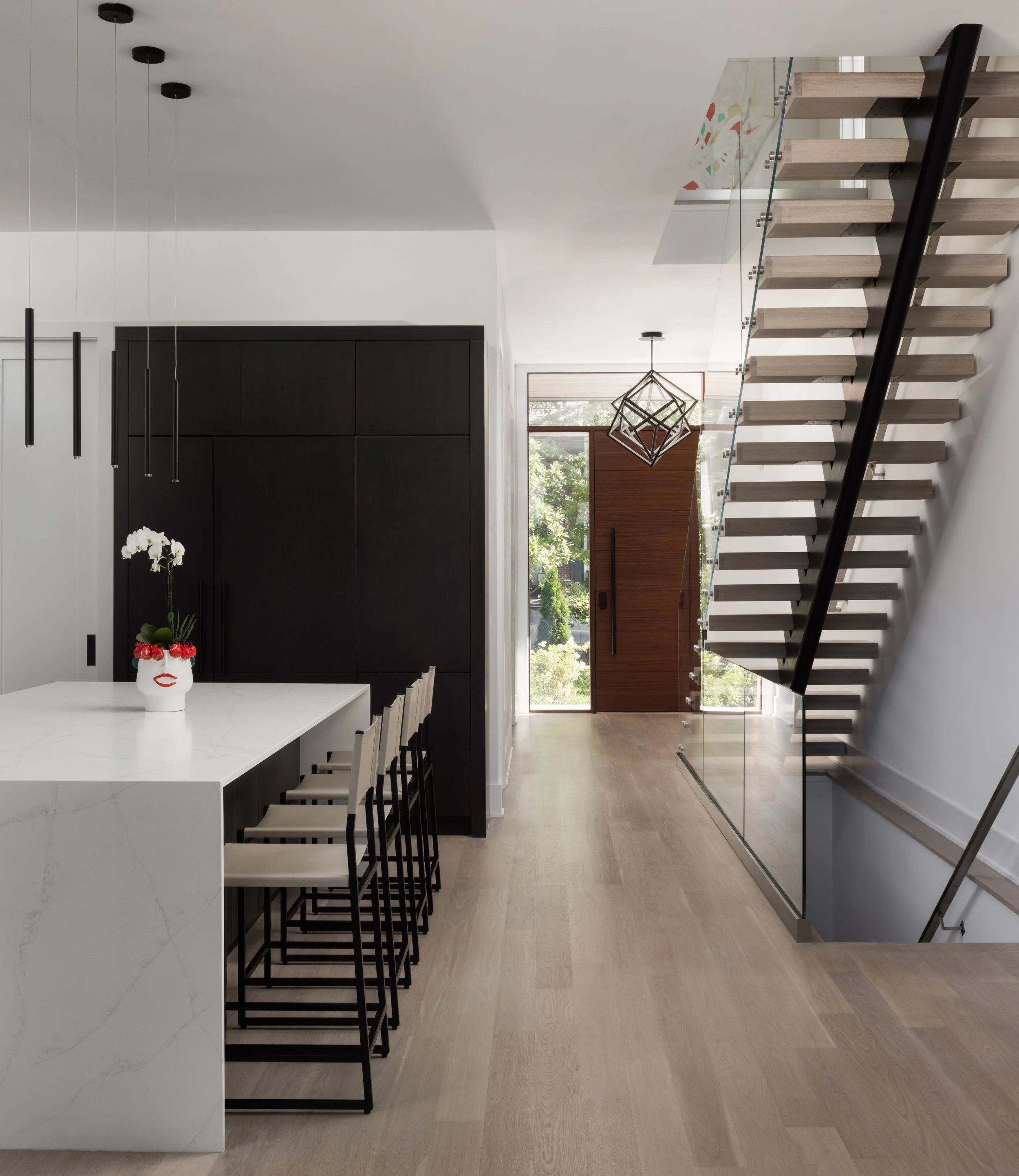Modern interior with a staircase, black cabinetry, a white table with chairs, a decorative face vase, pendant lights, and large windows with greenery outside.
