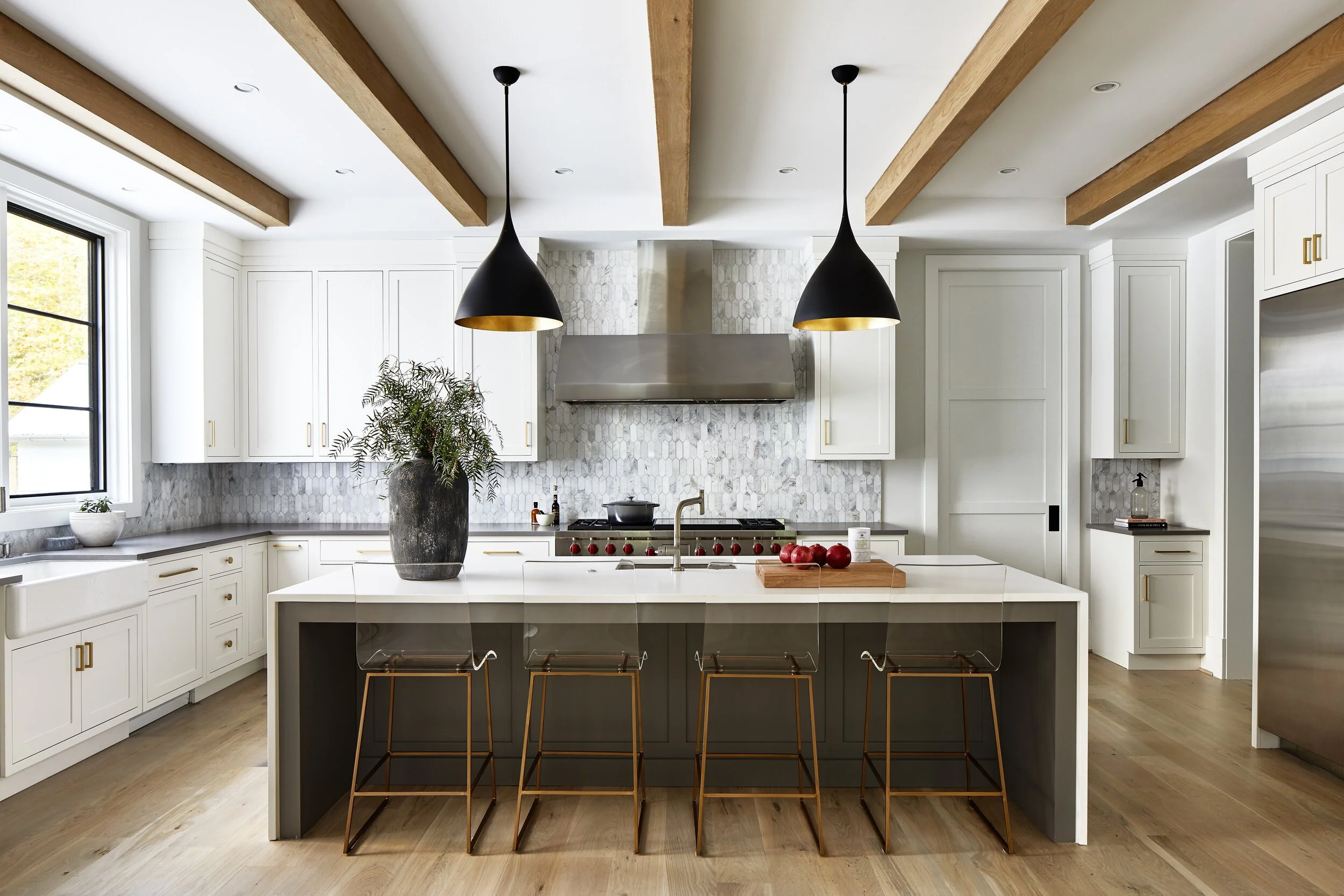 Modern white kitchen with a large island, black pendant lights with gold interior, and a vase with green foliage on the island. White cabinetry with gold handles, a stainless steel range hood, and a window with black trim are visible.