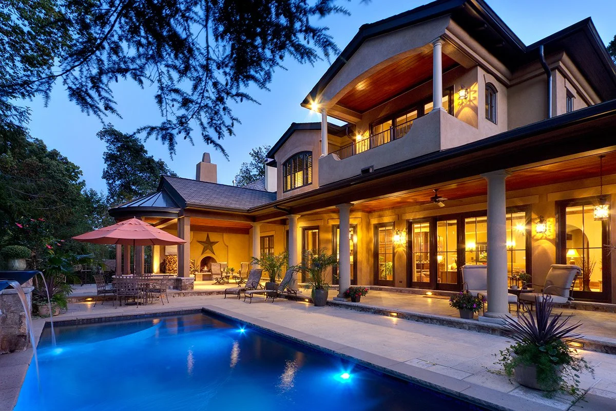 A two-story house with a swimming pool in the backyard during twilight. The house has large windows, a balcony on the upper floor, and outdoor lighting.