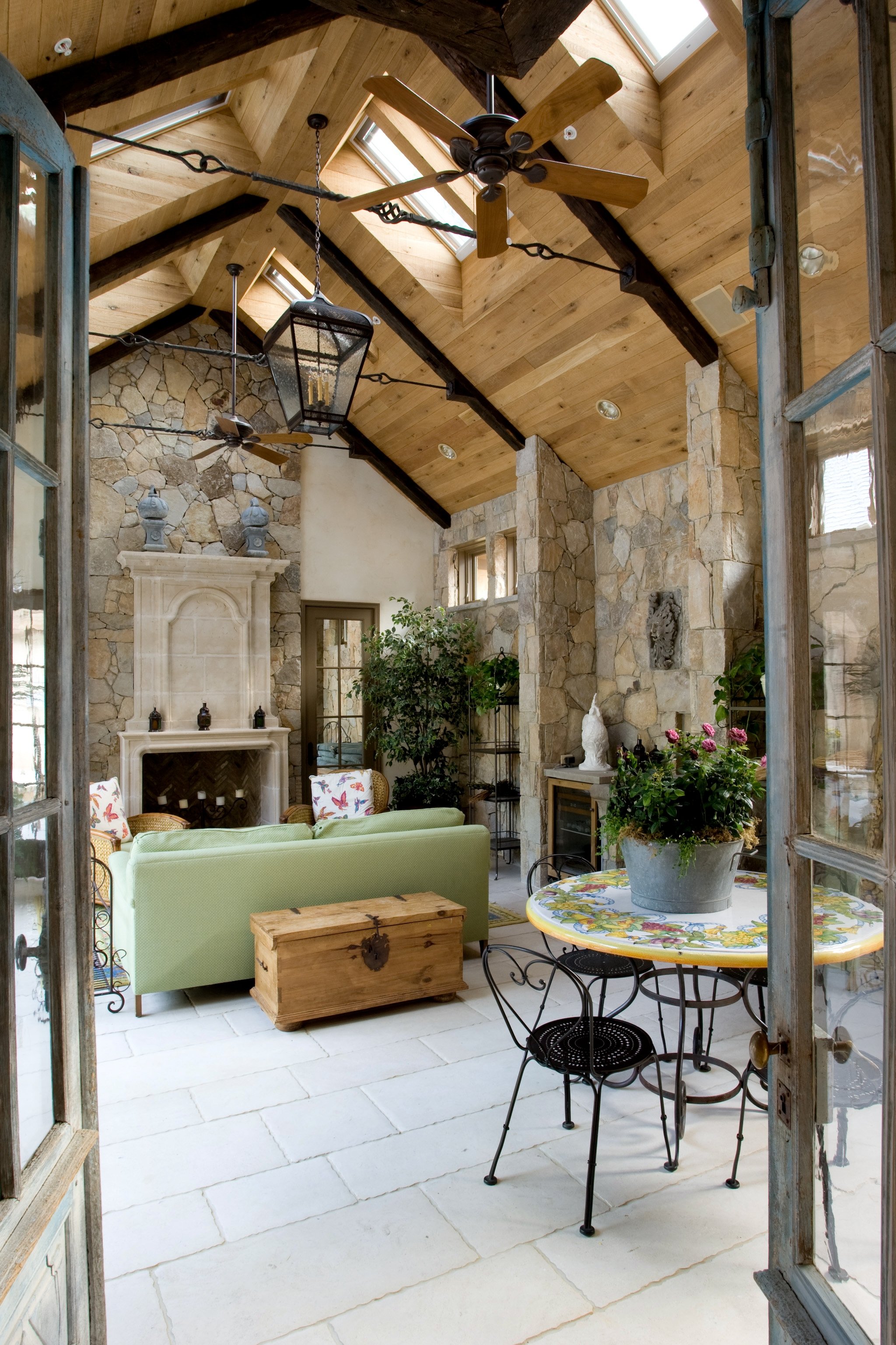 A sunlit interior living space with a vaulted wooden ceiling and skylights, stone walls, a fireplace, and various chairs and tables, including a green couch, a colorful round table with a potted plant, and a small wooden chest.