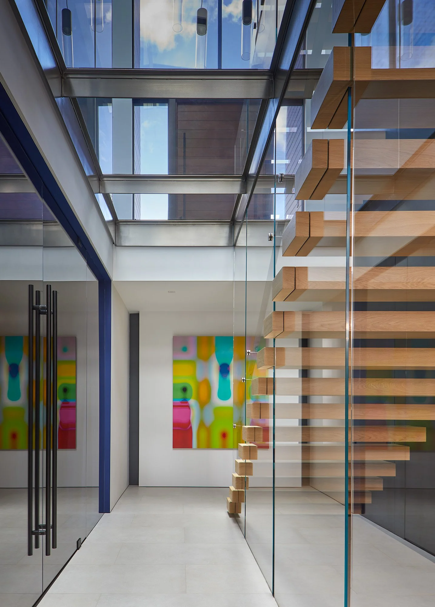 Modern interior space with glass walls, wooden stairs, colorful abstract art on the walls, and a skylight with clouds and blue sky visible.
