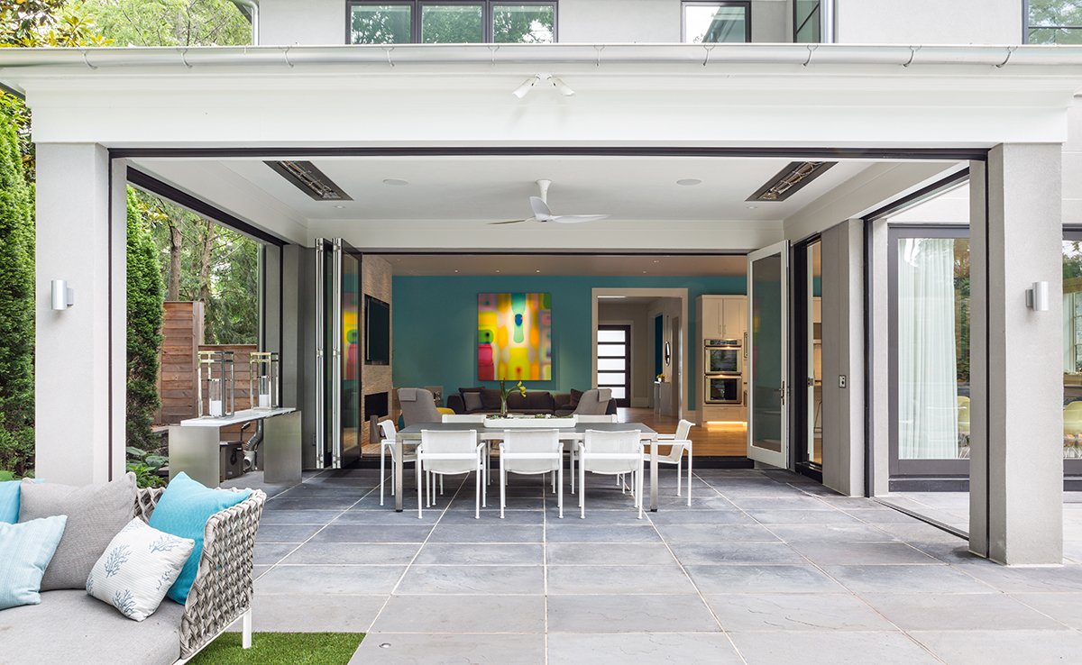 Open-concept outdoor patio and indoor living space with large glass sliding doors, a dining table with white chairs, colorful art on blue wall, and modern kitchen in the background.