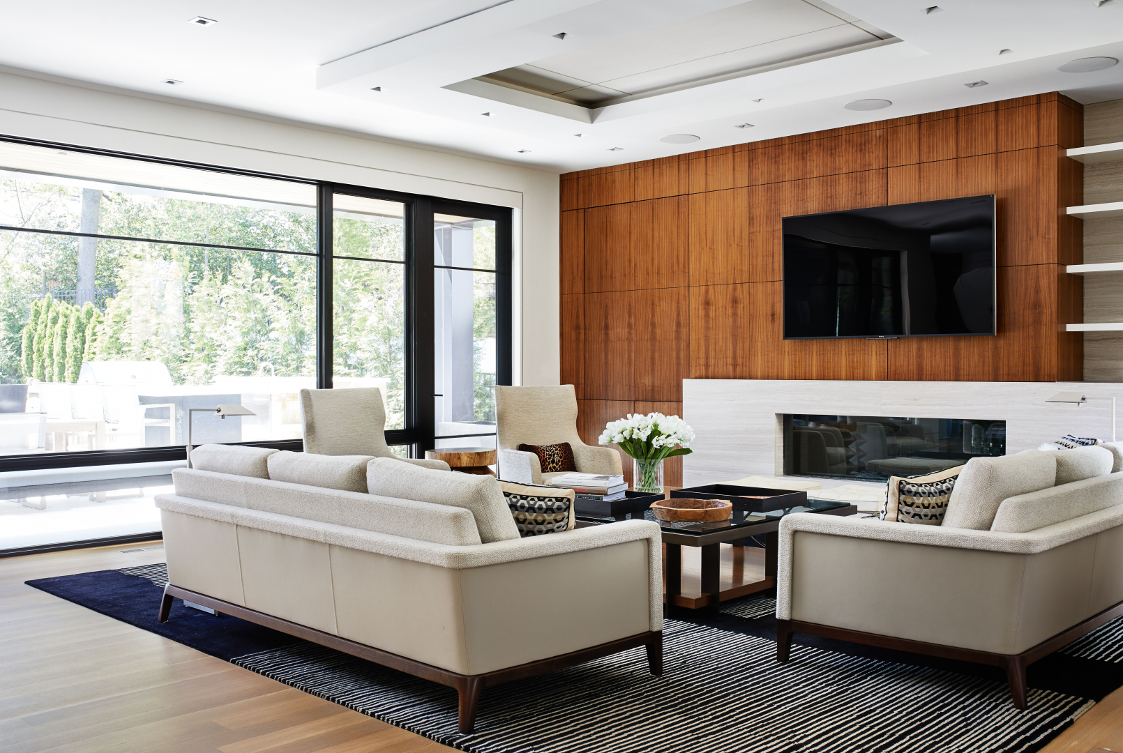 Living room with beige couches, a glass coffee table, and a black rug in front of large windows with outdoor greenery. A wooden accent wall with a mounted flat-screen TV and a modern fireplace below it. White and beige decor with a vase of white flowers on the coffee table.