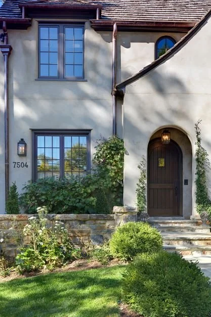 Front exterior of a beige house with dark gray trim and a dark wooden front door, surrounded by greenery and stone steps, with the house number 7504 visible.