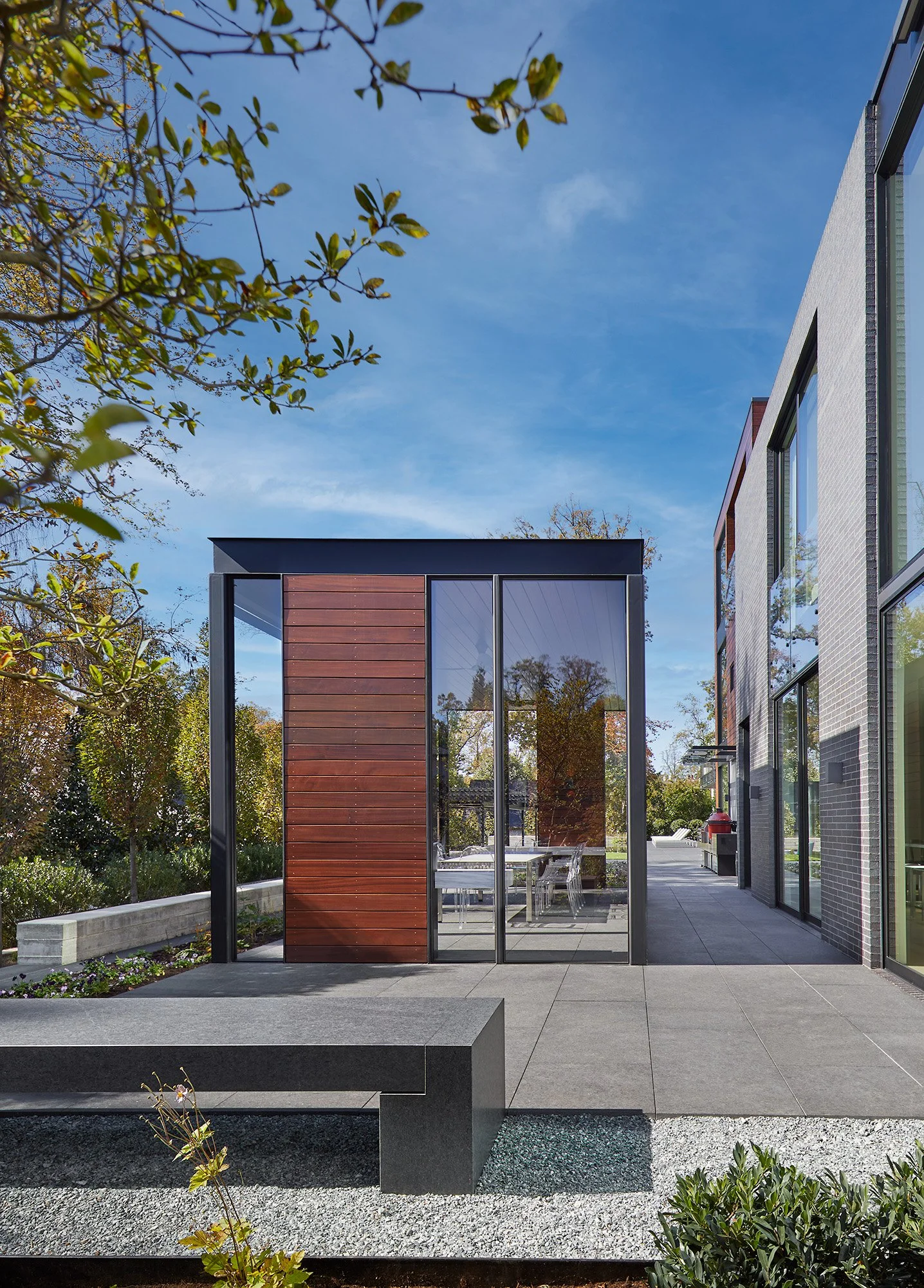 Modern residential outdoor patio with glass-enclosed wooden structure, concrete flooring, surrounding greenery, and a building with large windows.