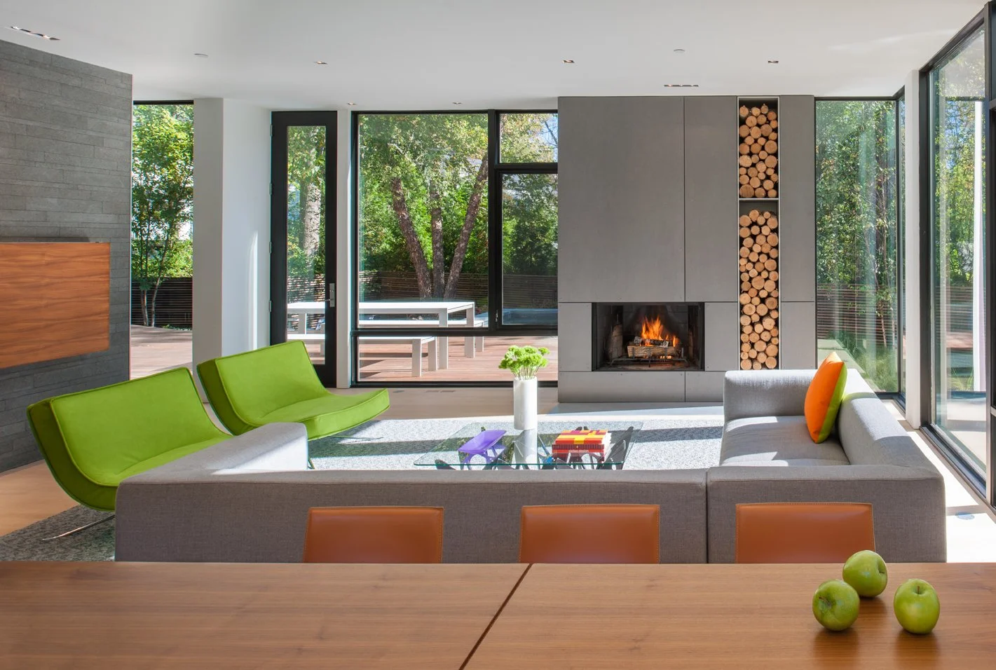 Modern living room with gray couch, bright green chairs, a fireplace, and large glass windows showing outdoor deck and trees.