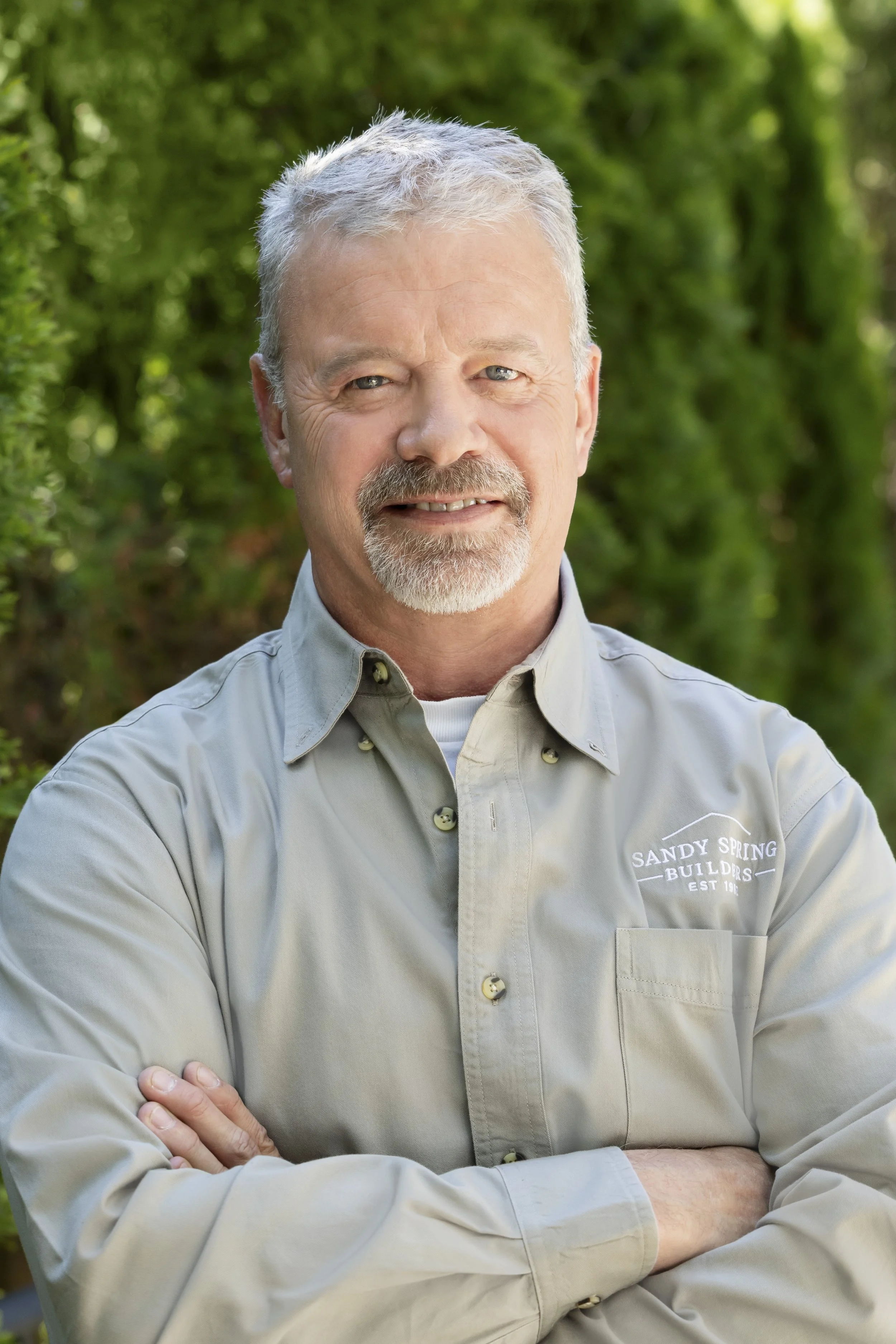 A middle-aged man with gray hair and a beard, wearing a light gray shirt with a logo that reads 'Sandy Spring Builders Est 1981', standing outdoors with crossed arms and a confident smile, with green trees in the background.