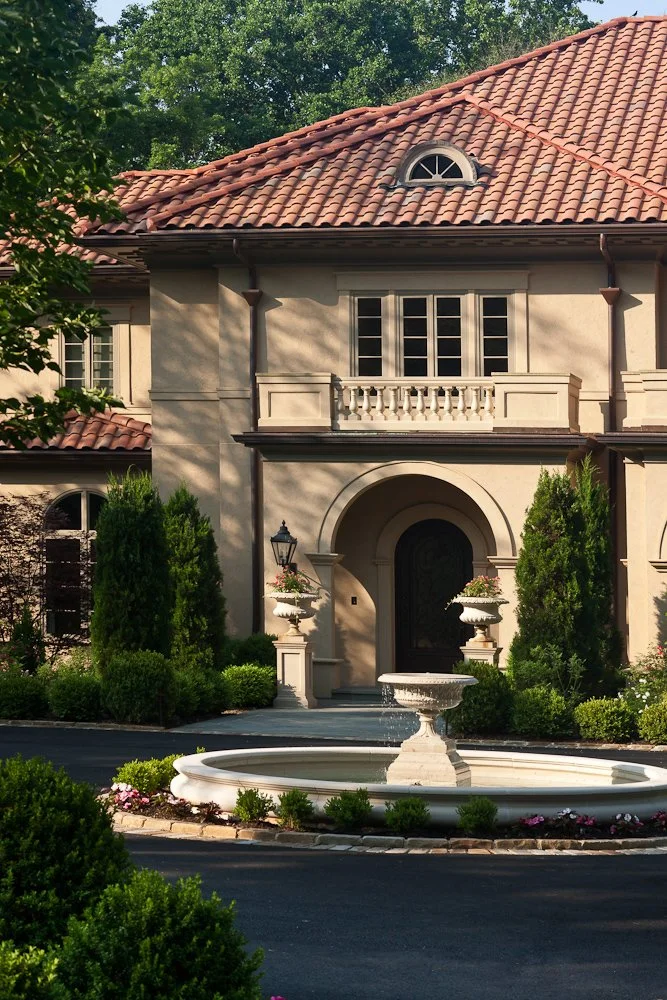 A large, elegant house with a tiled red roof, arched windows, and a grand entrance framed by an archway. The front yard features a round fountain, manicured bushes, and tall trees.