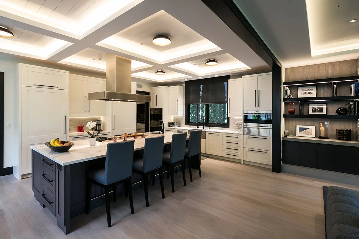 Modern kitchen with white cabinets, a large island with a white countertop, four gray bar stools, built-in appliances, black window shades, and a black and wood shelving unit with decorative items.