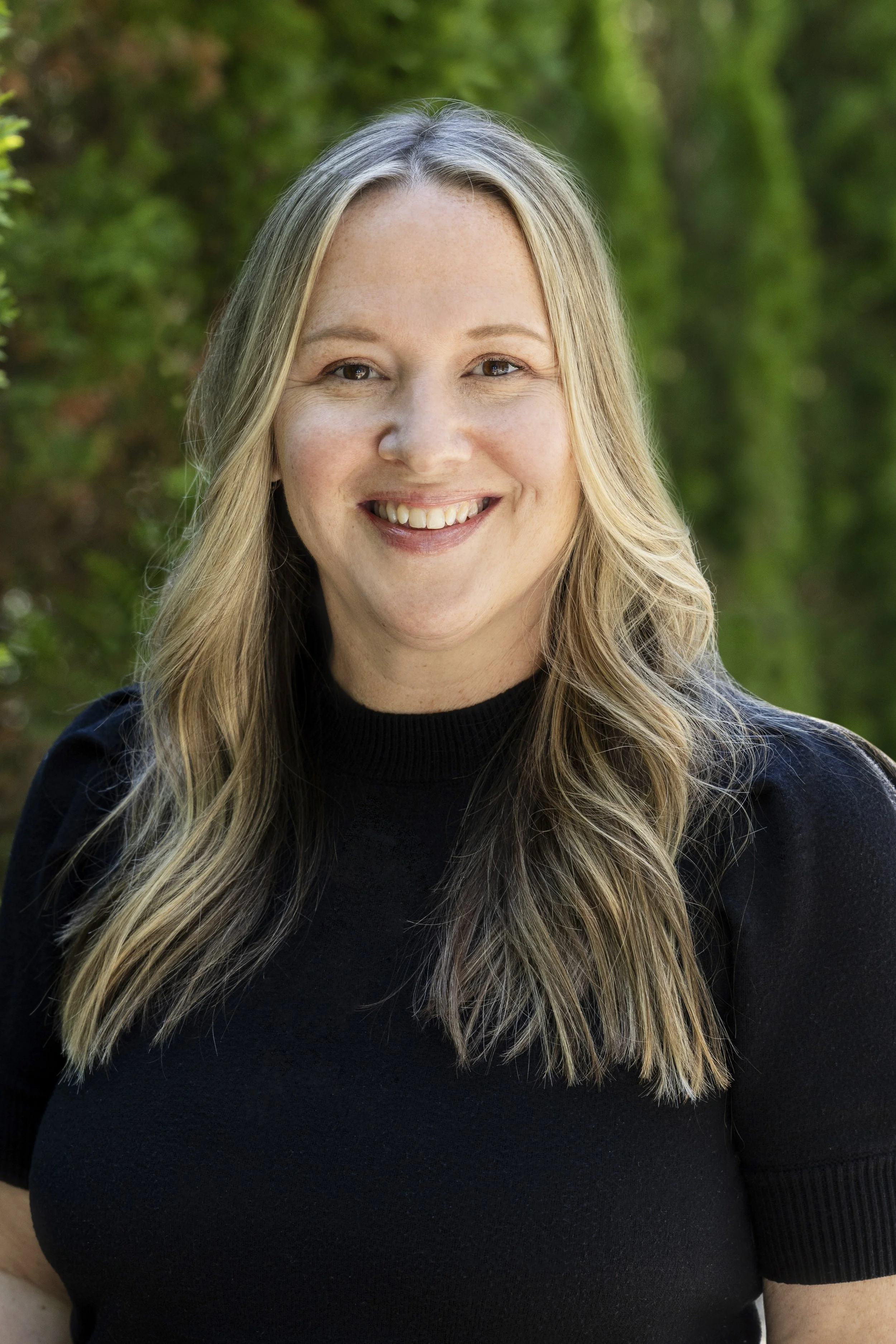 Close-up of a smiling woman with long, wavy blonde hair, wearing a black top, standing outdoors with green foliage background.