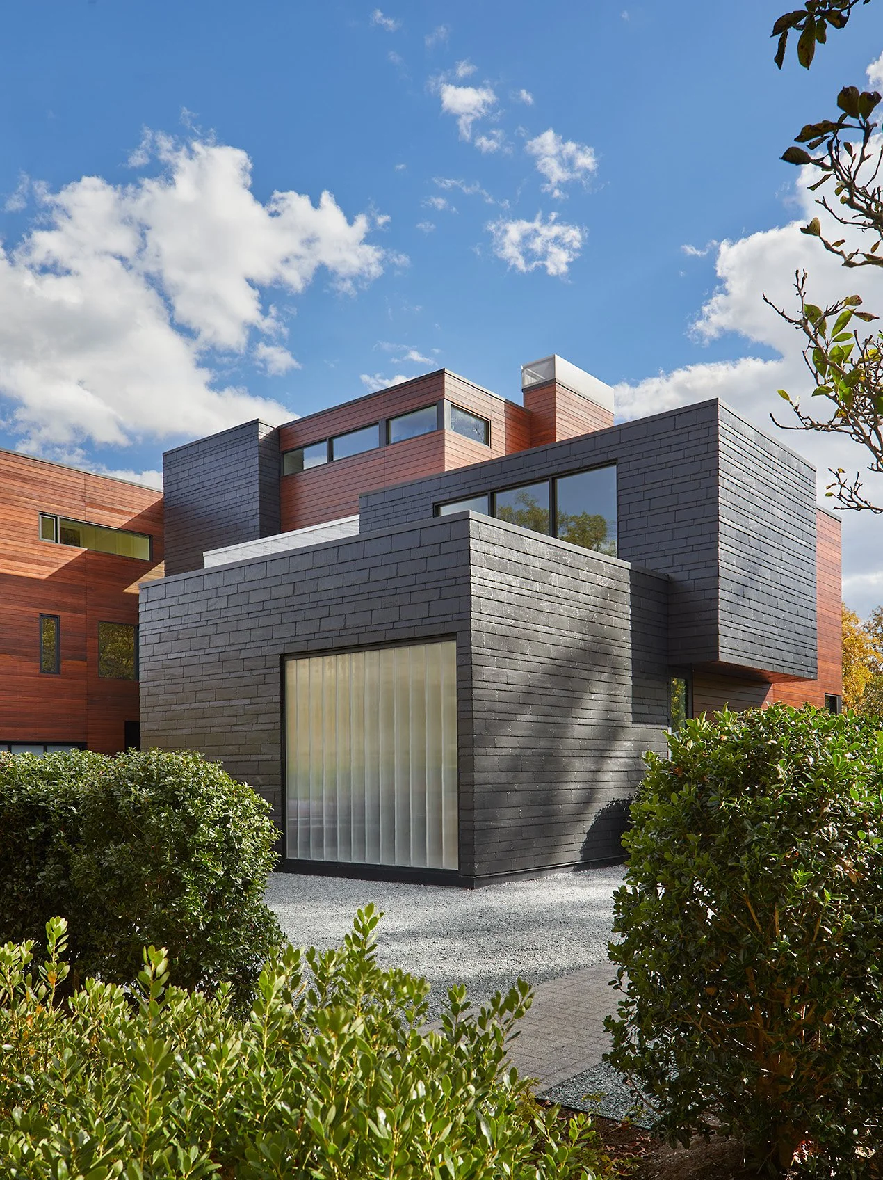 Modern multi-story house with black brick and wooden siding exterior, large windows, and a gravel pathway surrounded by greenery under a blue sky with scattered clouds.