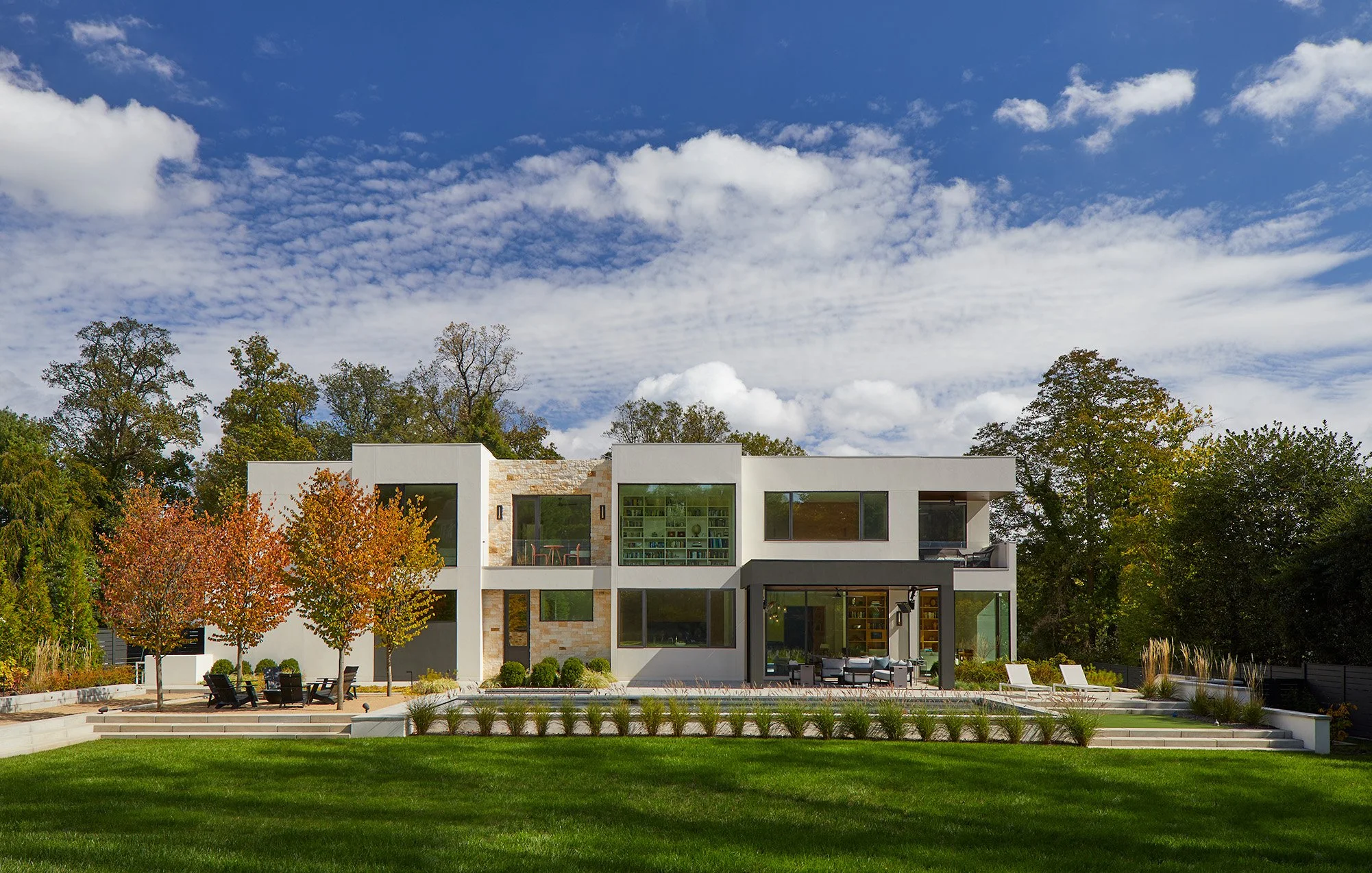 Modern two-story house with large windows, surrounded by trees with colorful autumn leaves, under a blue sky with clouds.