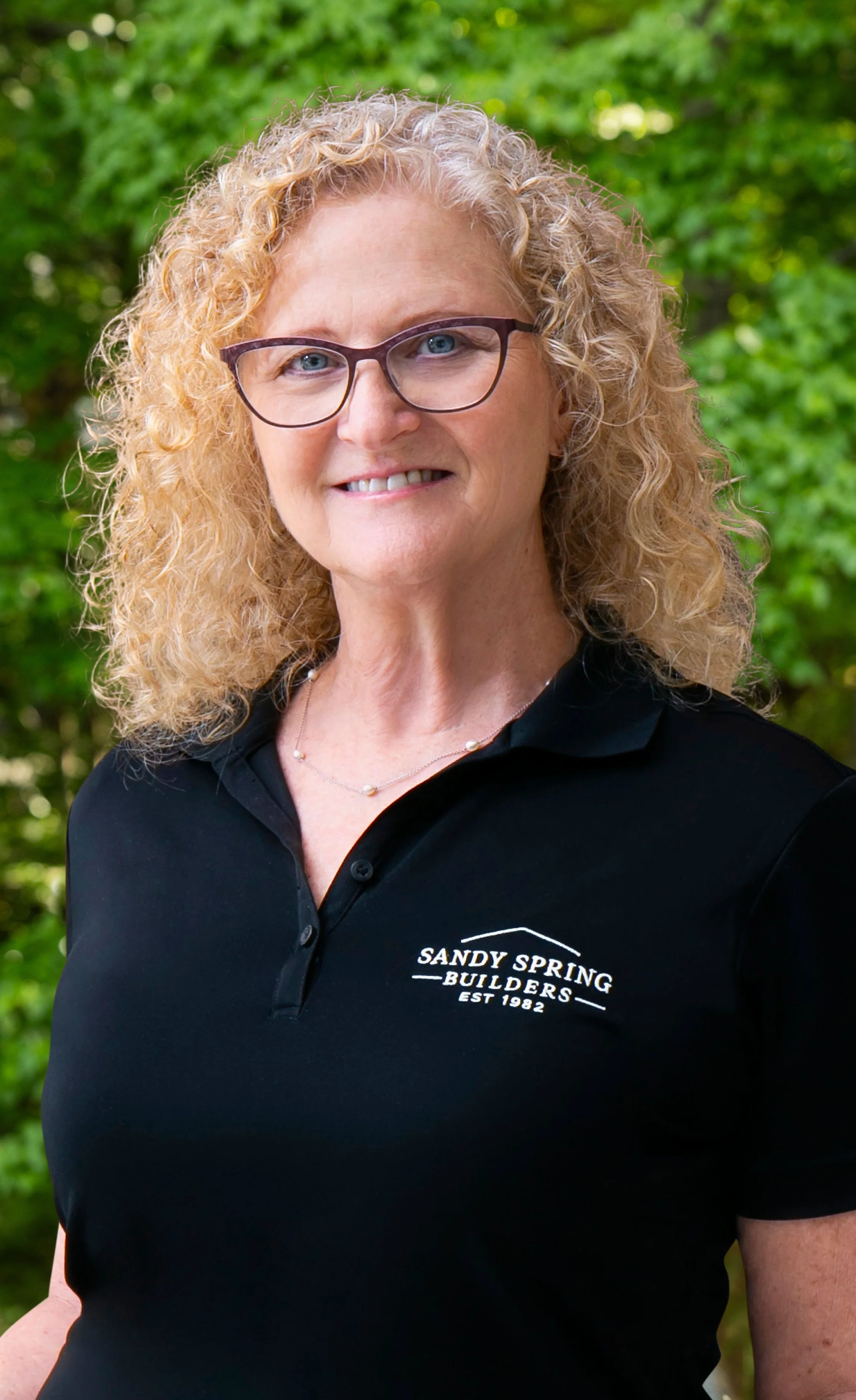 A woman with curly blonde hair, wearing glasses and a black polo shirt with a logo that reads 'Sandy Spring Builders, Est 1982,' standing outdoors with green foliage in the background.
