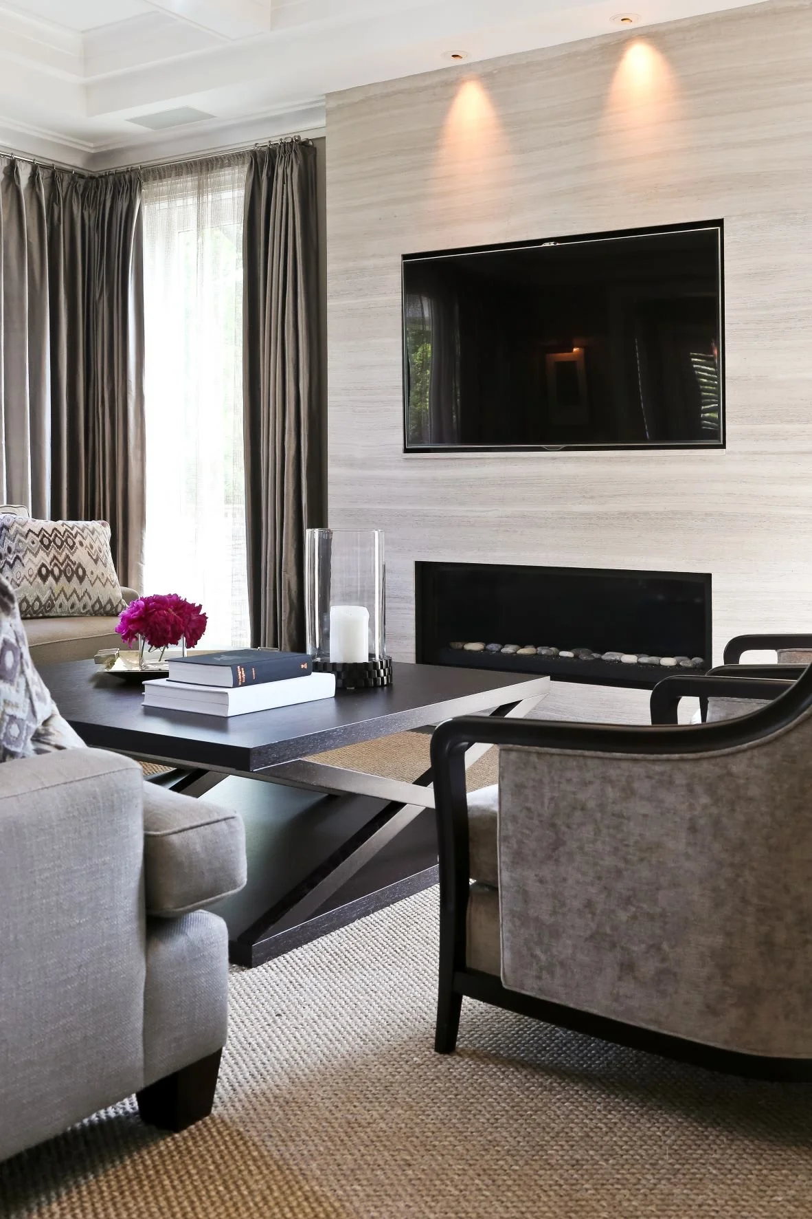 Modern living room with a wall-mounted TV, a fireplace, a black coffee table with books, pink flowers, a glass candle holder, and various chairs and curtains.