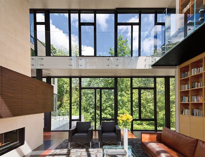 Modern living room with large glass windows, black chairs, a brown sofa, bookshelves, a fireplace, and a view of green trees outside.