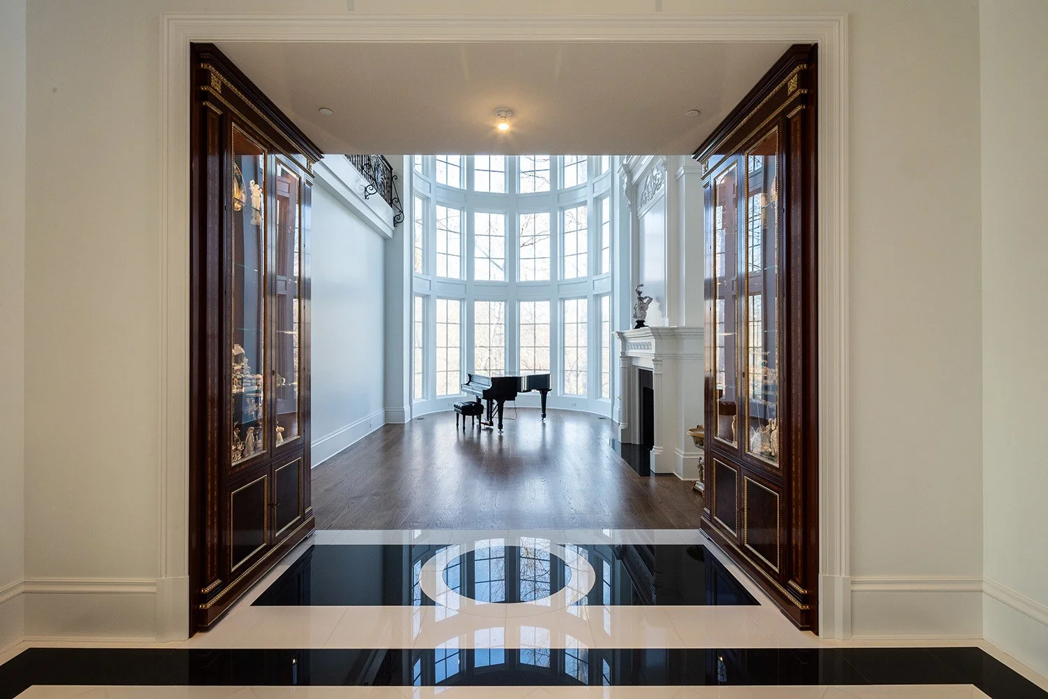 An elegant room with large bay windows, a grand piano, a fireplace with a sculpture on the mantel, and built-in wooden display cabinets.