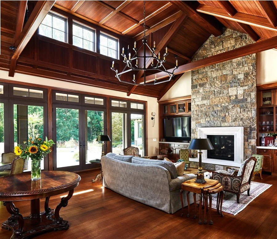 Living room with high wooden ceilings, a chandelier, stone fireplace, large glass doors, and a seating area with a sofa, armchairs, and lamps. There are windows letting in natural light and a vase with sunflowers on a round wooden table.