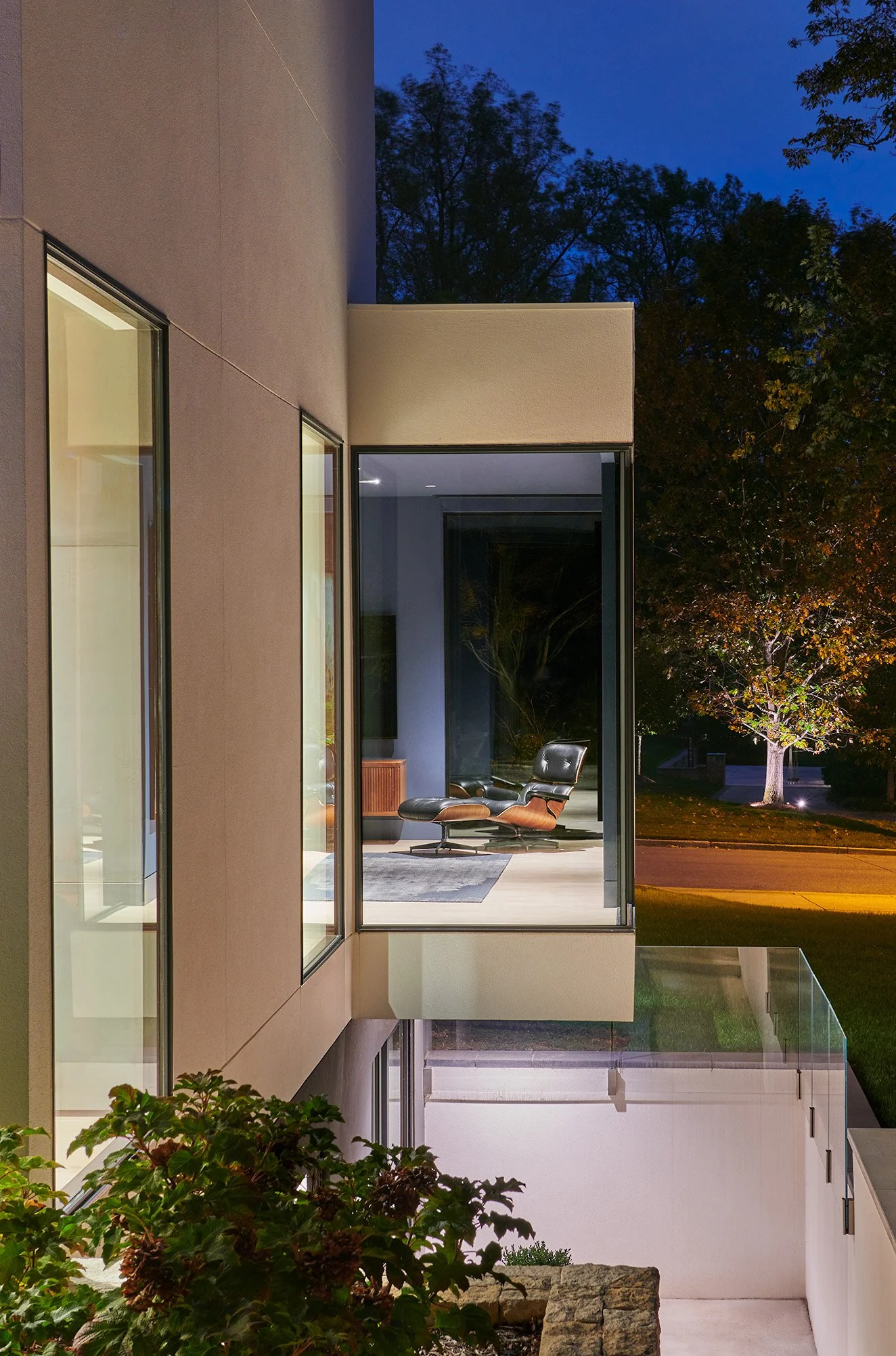 Modern house exterior at dusk with large glass windows, visible interior seating area with a black leather lounge chair and a gray rug, trees and a lawn outside, balcony with glass railing, night sky above.