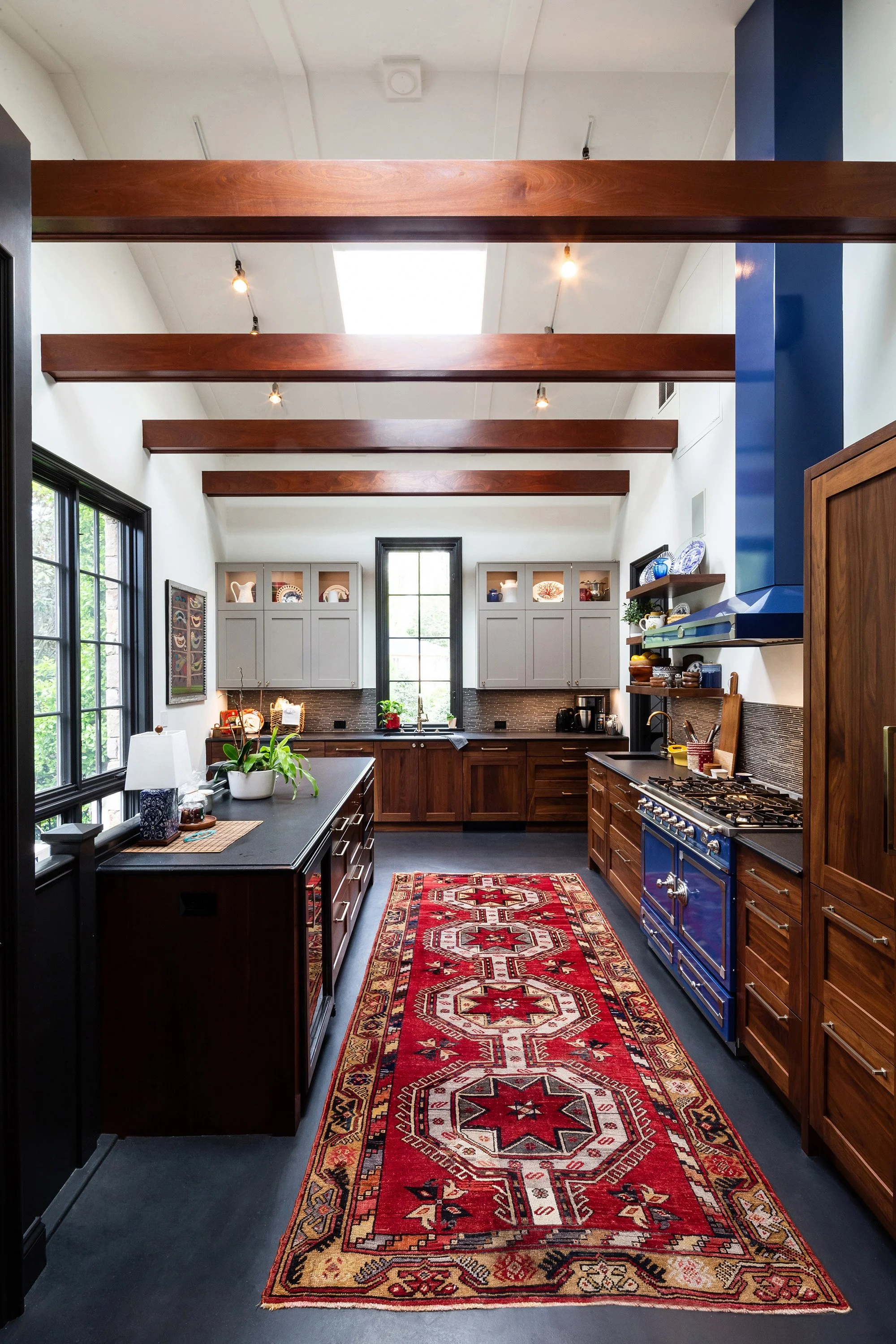 A kitchen with dark wooden cabinets, a blue stove, and a colorful red patterned runner rug on the black floor. The ceiling has exposed wooden beams with recessed lighting. Large windows on the left let in natural light.