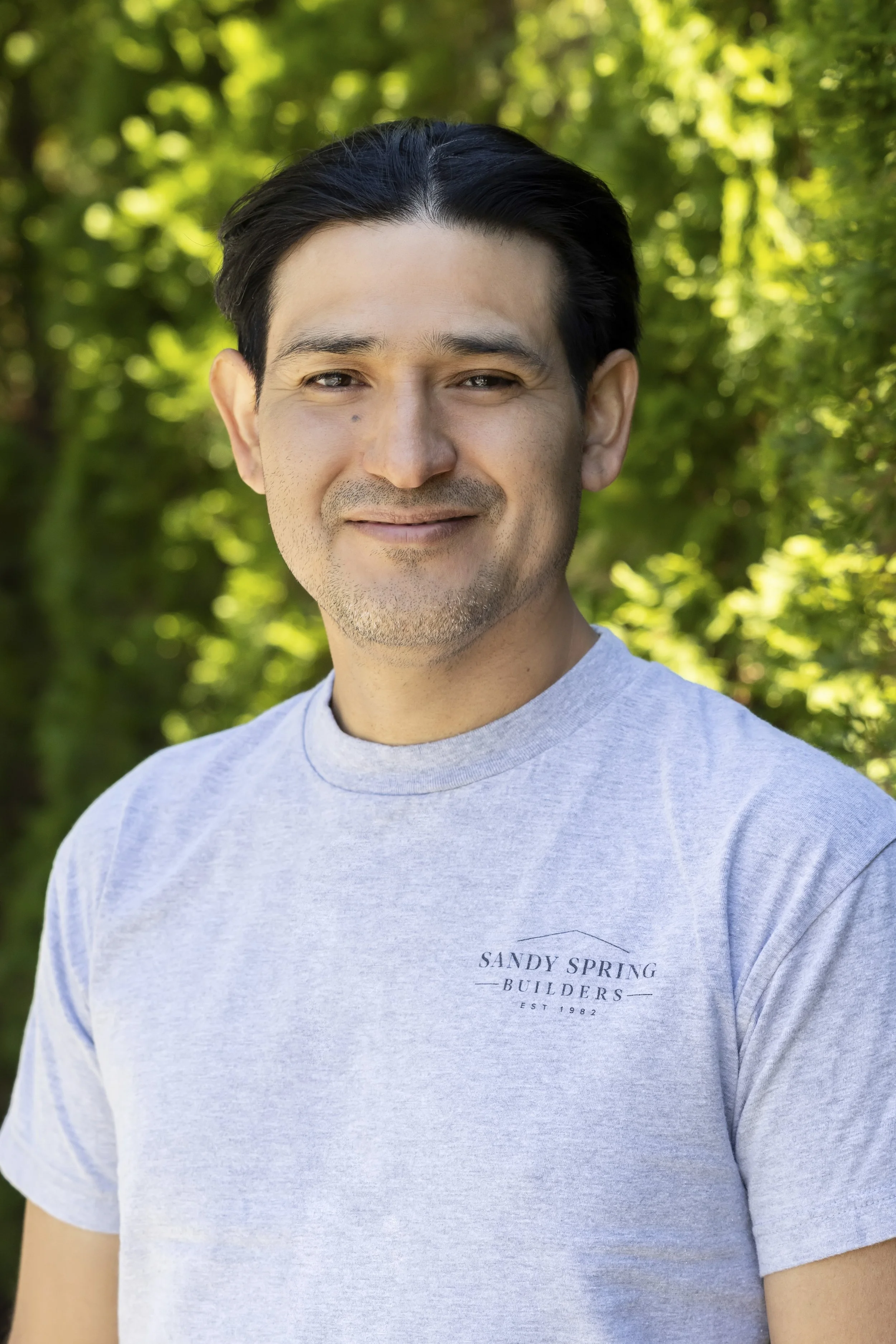 Close-up of a man with dark hair, light skin, wearing a light gray T-shirt with 'SANDY SPRING BUILDERS' printed on it, outdoors with green trees and sunlight in the background.