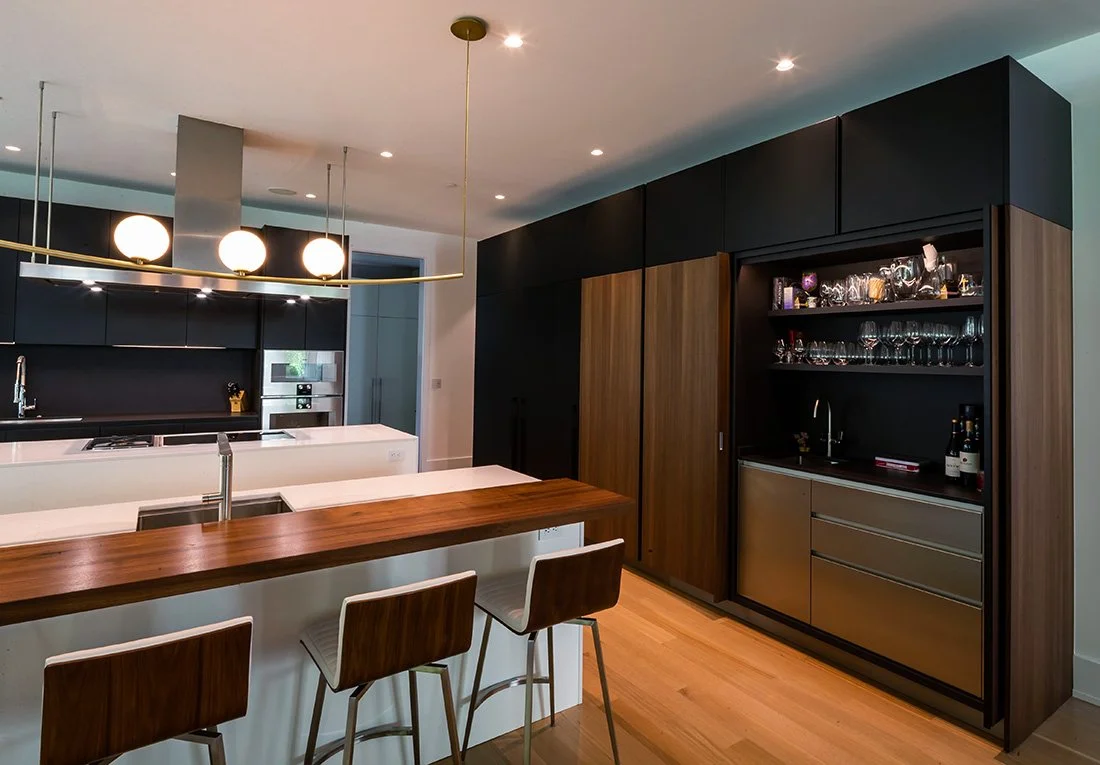 Modern kitchen with a white island, wooden bar counter, black cabinets, and a built-in bar area with glassware and wine bottles.