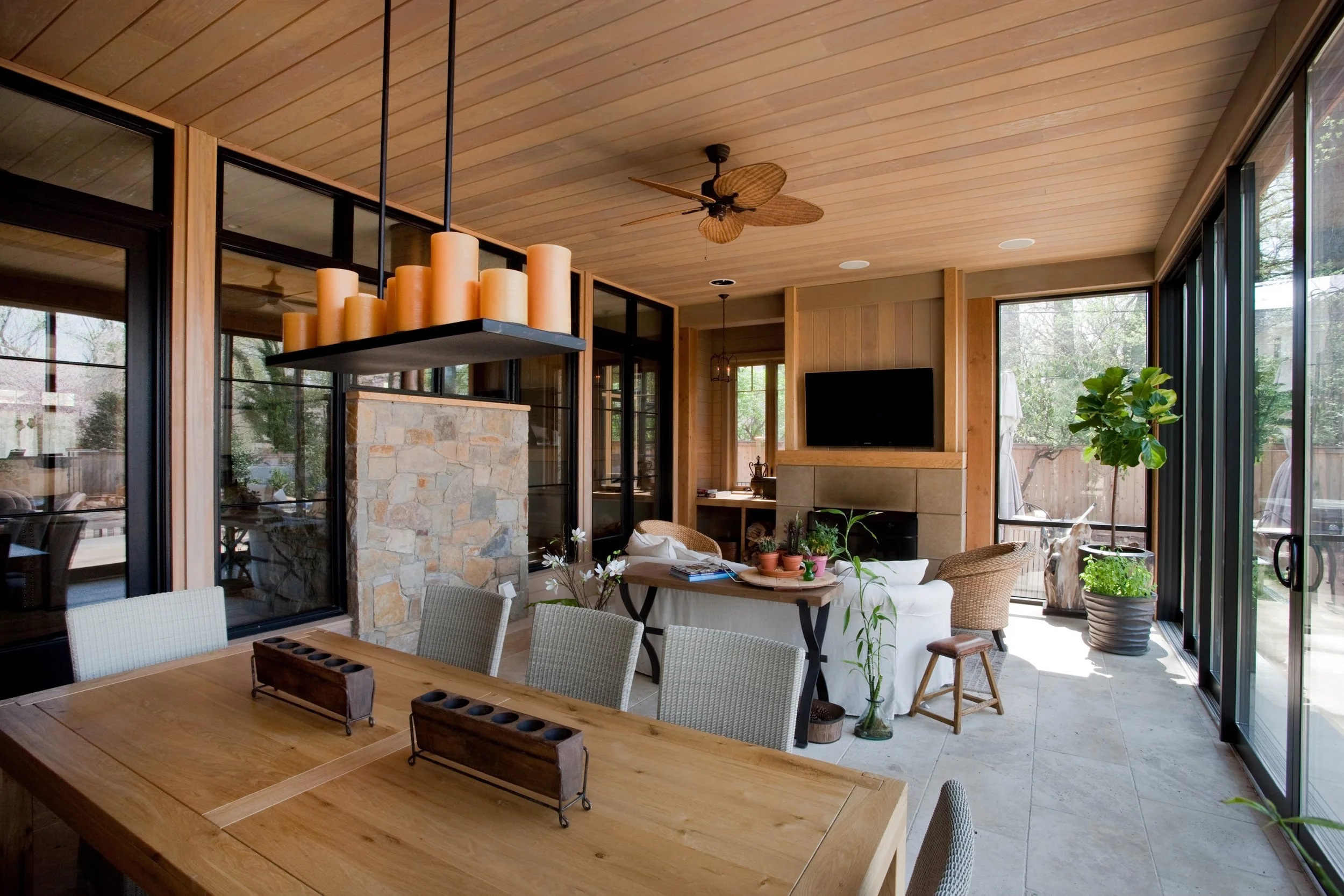A bright living room with large glass windows and a wooden ceiling, featuring a ceiling fan, a stone fireplace, a flat-screen TV, and various plants and furniture.