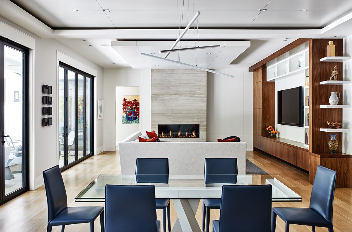 Modern living room with glass dining table, black chairs, white sofa, fireplace, and built-in wooden entertainment center with a TV and decorative items, sunlit by large sliding glass doors.