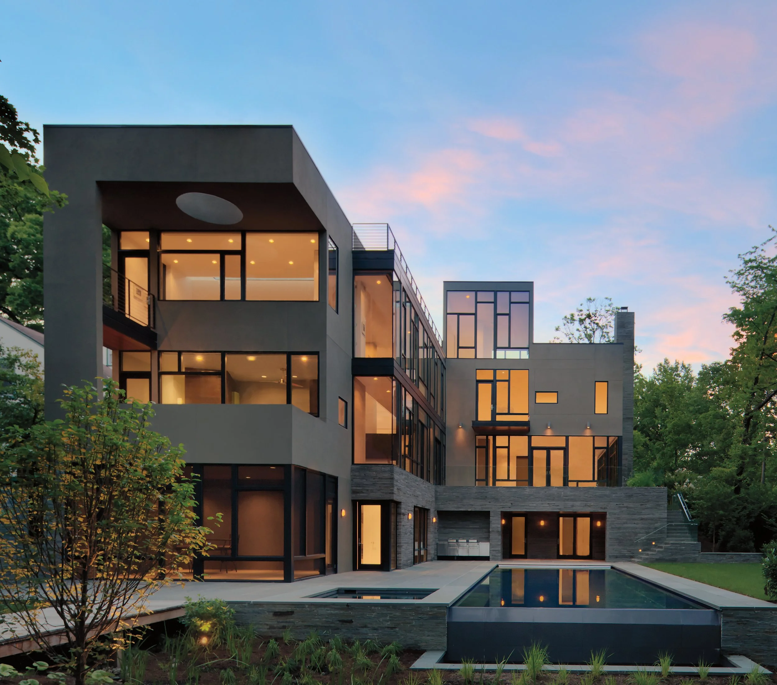Modern multi-story house with large glass windows, illuminated from inside, surrounded by trees and a pool area, during dusk or evening.