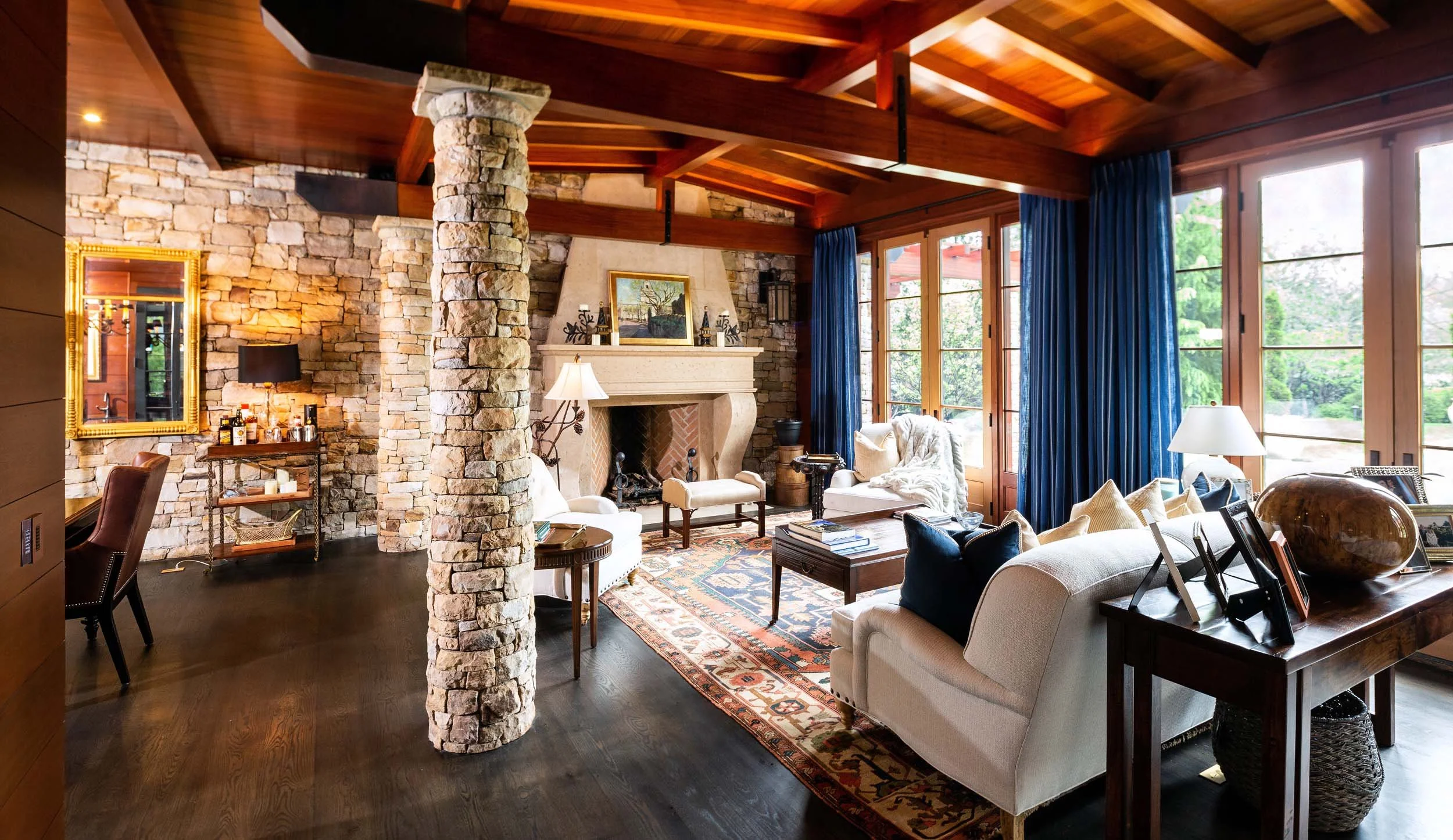 A cozy living room featuring a stone fireplace, wooden ceiling beams, large windows with blue curtains, and a variety of comfortable furniture and decorative items.