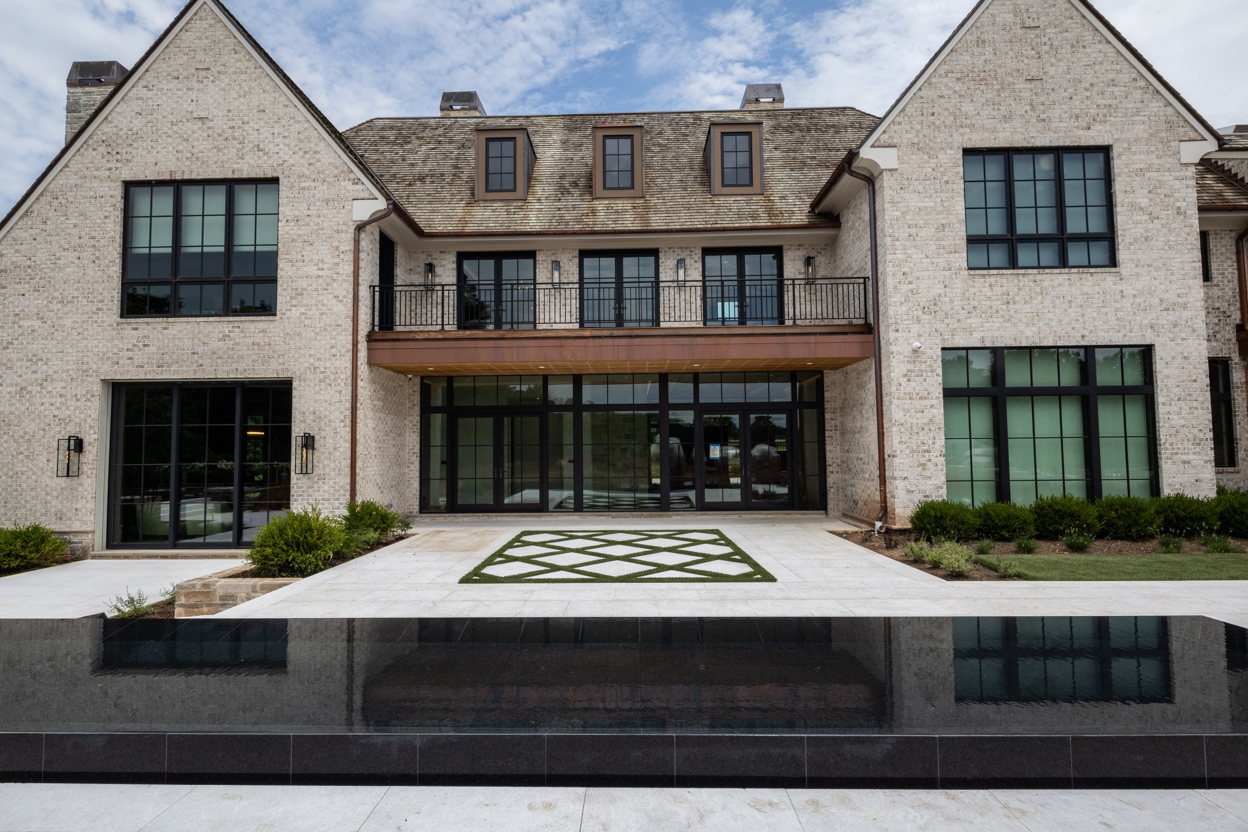 Exterior view of a modern brick house with large windows, a balcony, and a finished backyard with a fountain, plants, and a patterned walkway.