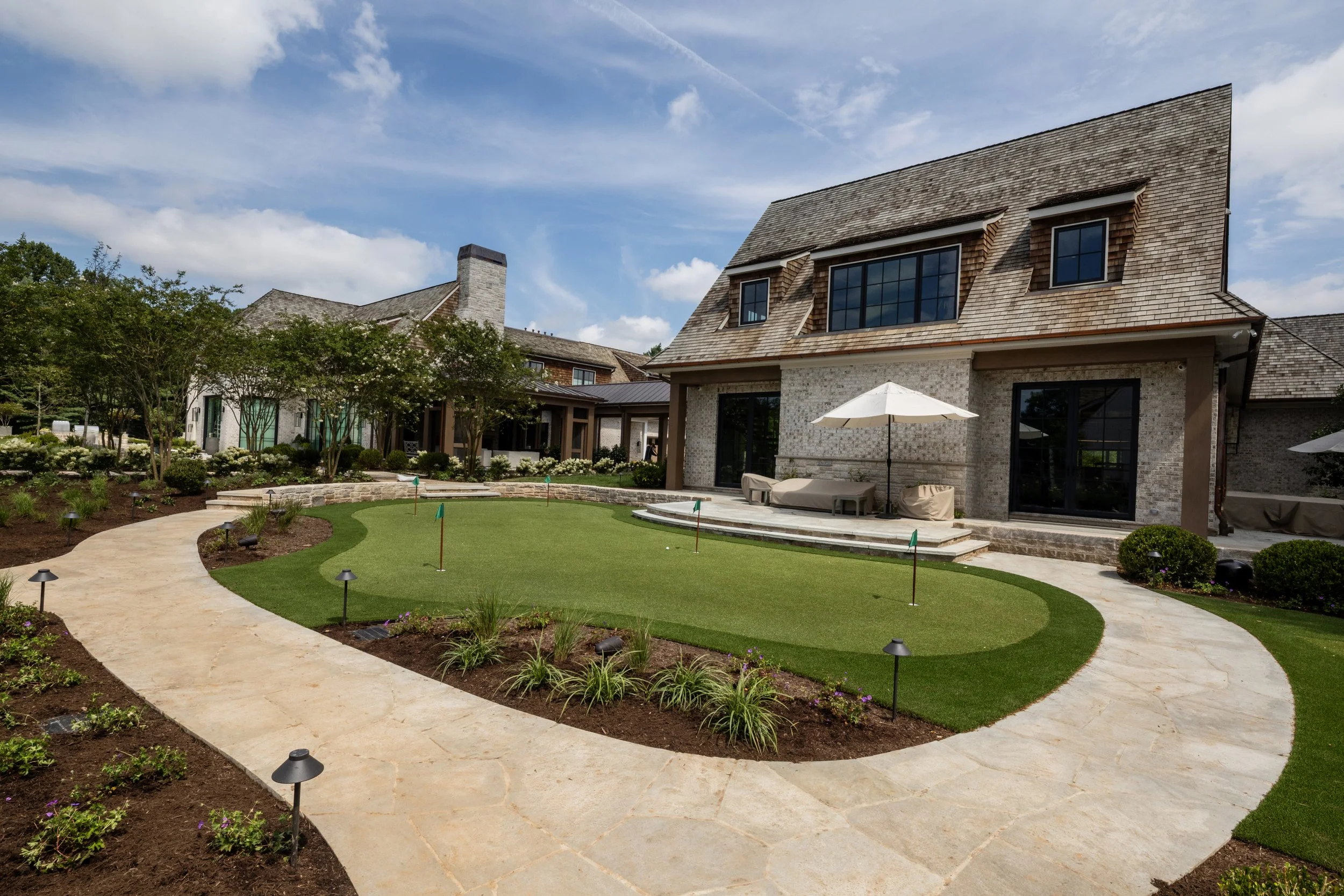 Front yard of a house with a curved stone pathway, green lawn, and landscaped flower beds, with a house featuring brick walls and a shingled roof in the background.