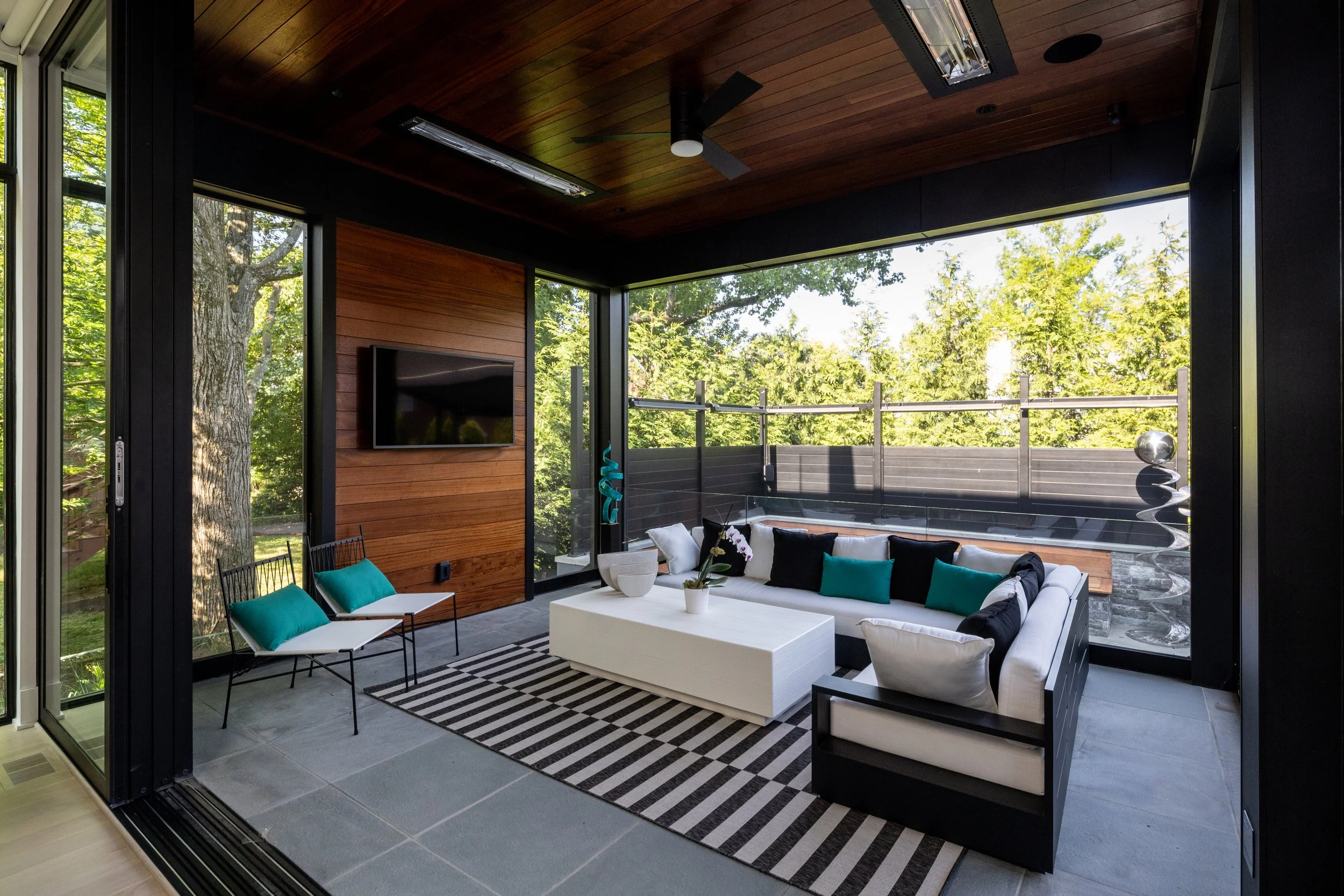 Modern indoor living room with large windows showing trees outside, wooden ceiling, ceiling fan, wall-mounted TV, white sofa with teal and black pillows, black chairs with teal cushions, and black and white striped rug.