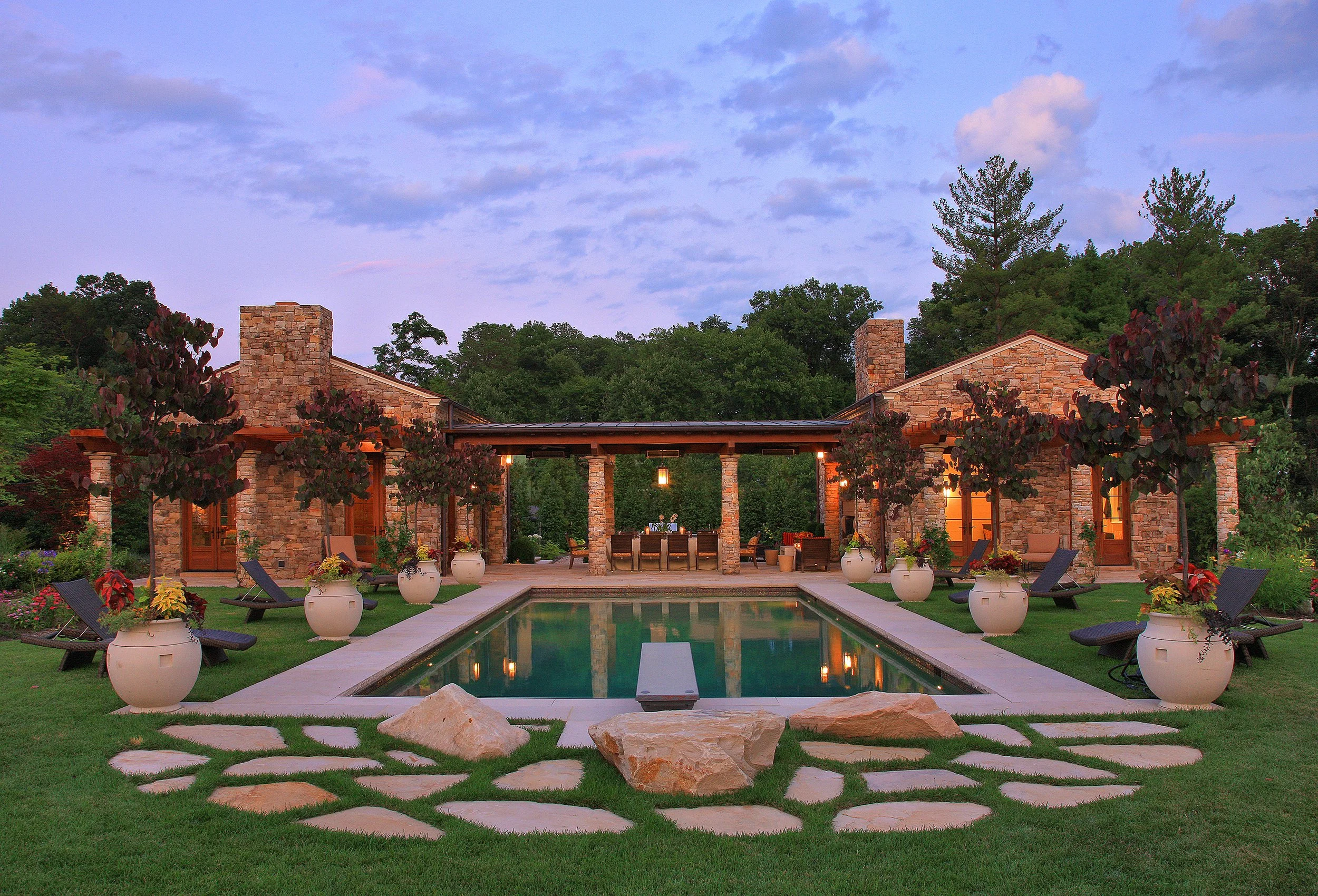 A backyard with a rectangular swimming pool, surrounded by potted plants, outdoor furniture, and large rocks, with a stone house and trees in the background during early evening.