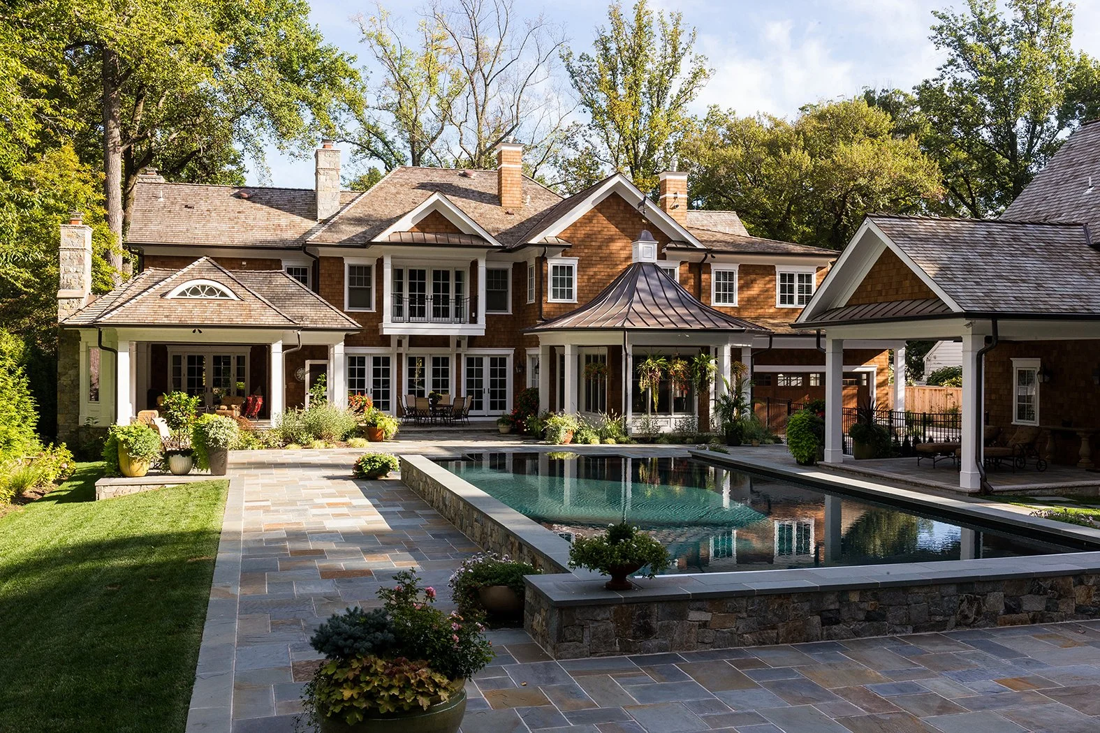 A luxurious brick house with multiple rooflines, glass doors, and a pool in the backyard. The yard has paving stones, potted plants, and outdoor furniture, surrounded by trees and greenery.