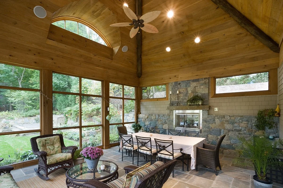 Sunroom with large windows, wooden ceiling, fireplace, dining table, and wicker furniture with cushions and plants.
