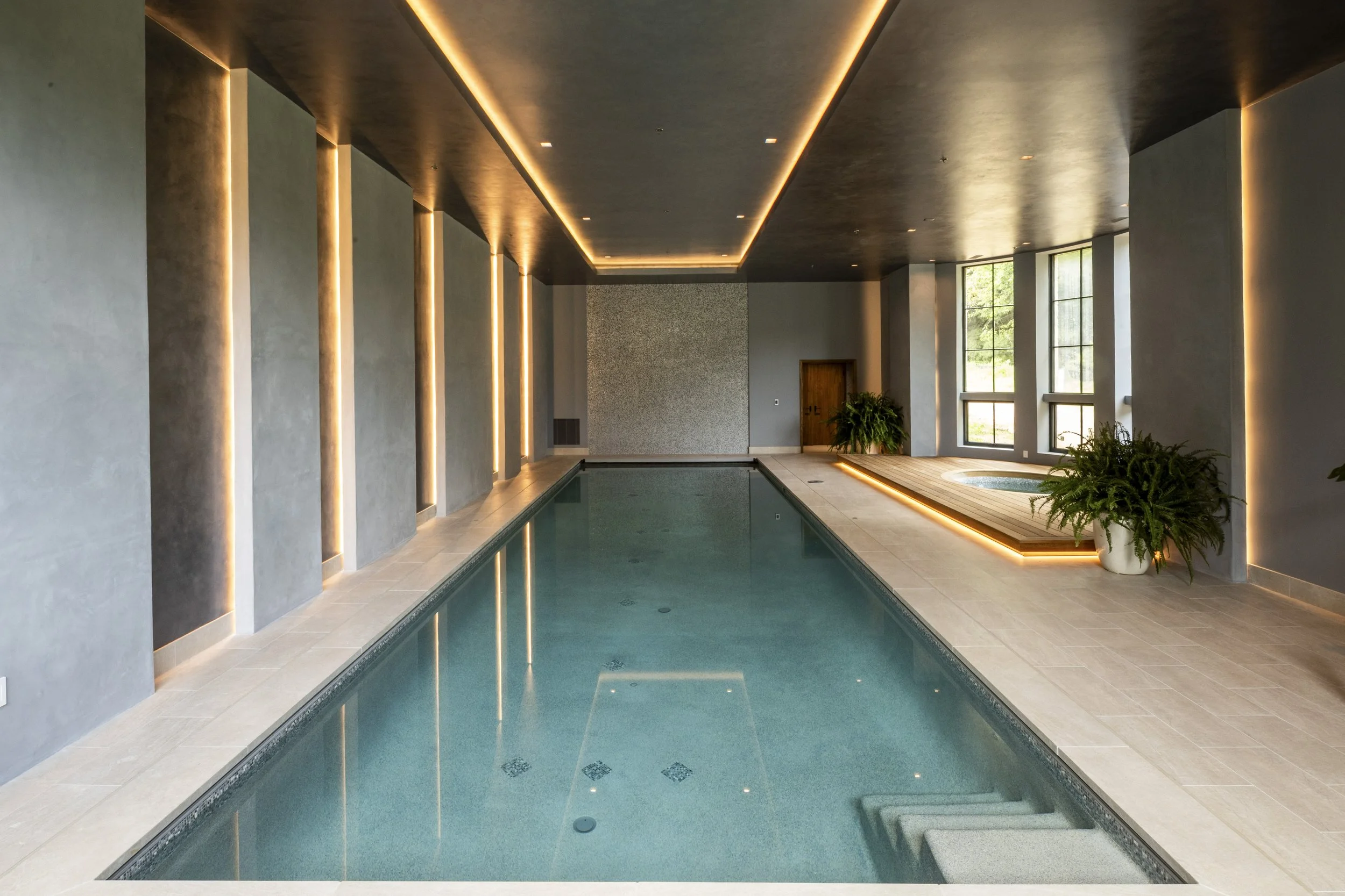 An indoor swimming pool with modern design, featuring blue water, beige tile flooring, and large windows with natural light. The ceiling has recessed lighting and embedded LED strip lights, with potted plants near the windows.