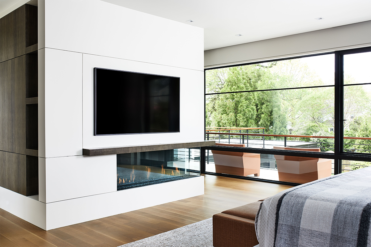 Modern bedroom with a flat-screen TV, a fireplace beneath the TV, a large window with a view of green trees, and a bed with a gray blanket.
