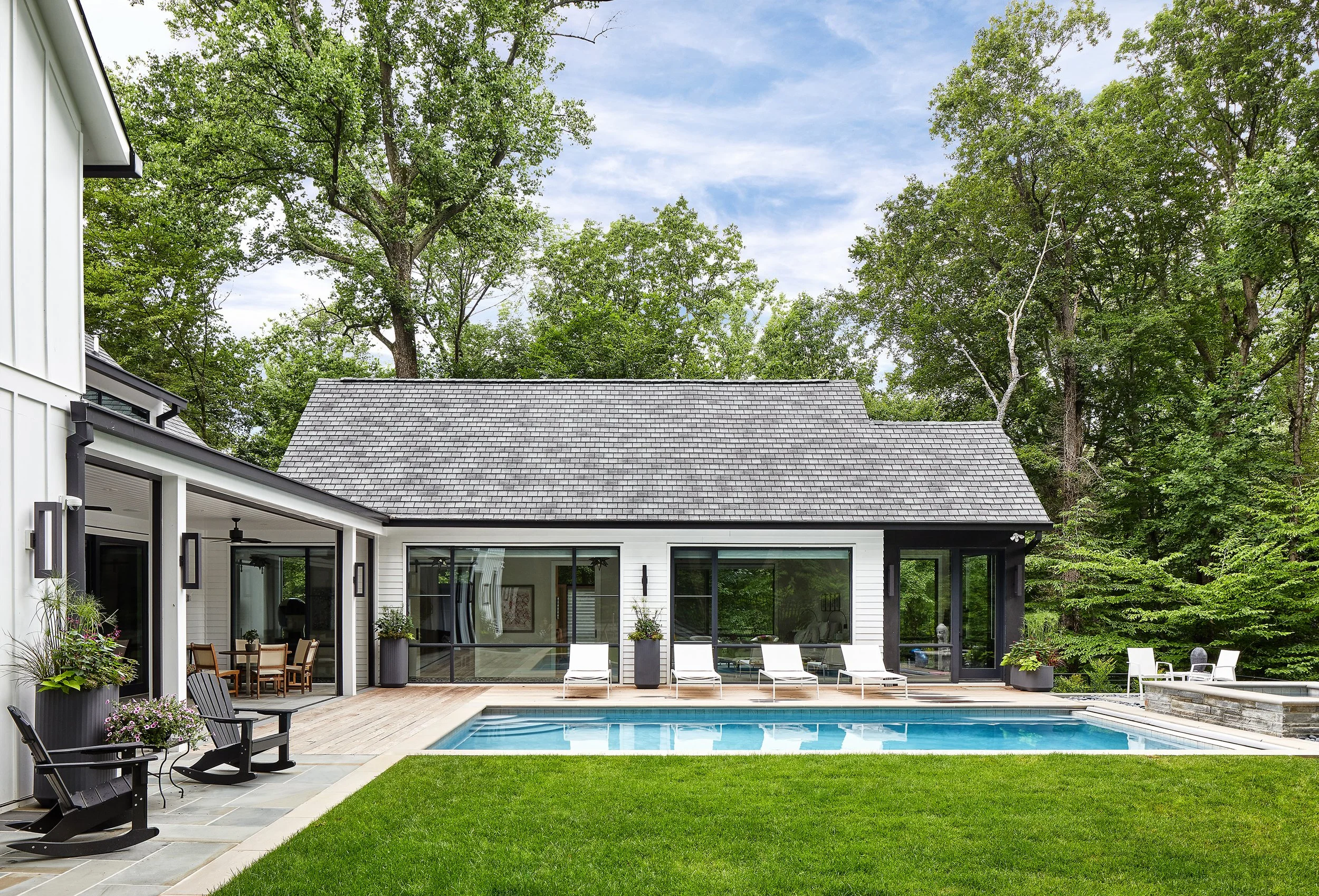 A modern house with large glass windows and a swimming pool in the backyard, surrounded by green grass, patio furniture, and tall trees in the background.