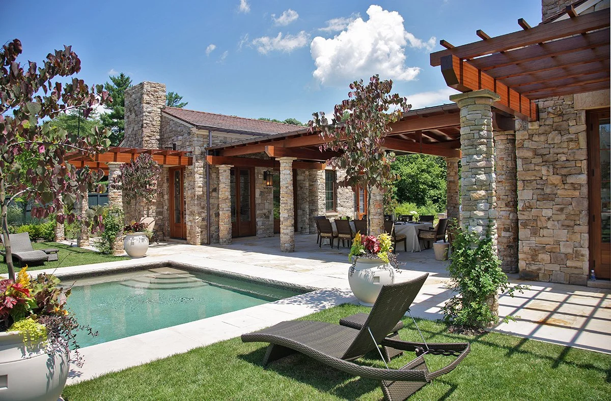 Backyard patio with a small pool, lounge chair, potted plants, and a covered dining area with stone columns and wooden beams, surrounded by greenery and trees under a partly cloudy sky.