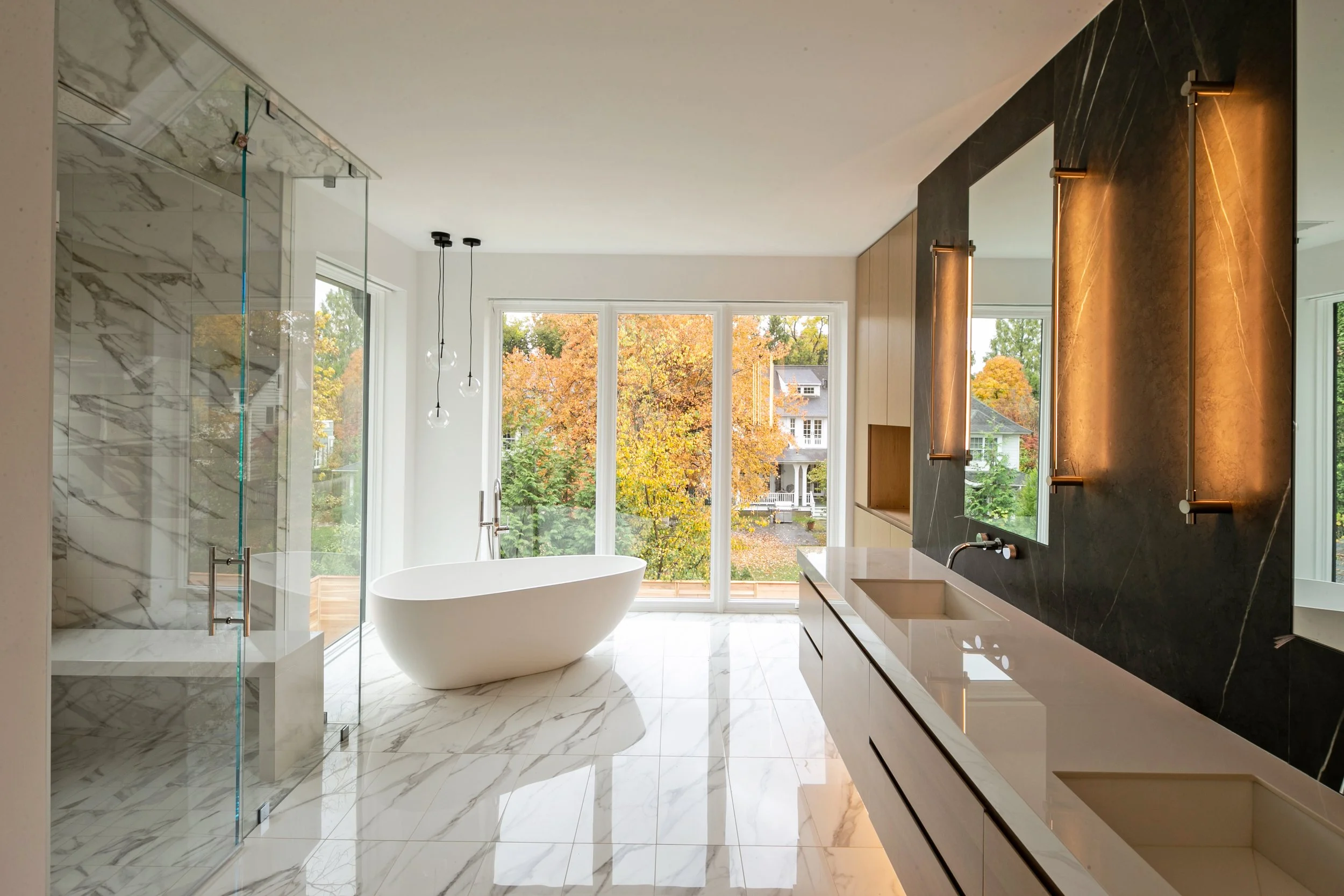 Modern bathroom with a freestanding bathtub, marble floor and walls, a glass-enclosed shower, and large windows showing autumn trees outside.