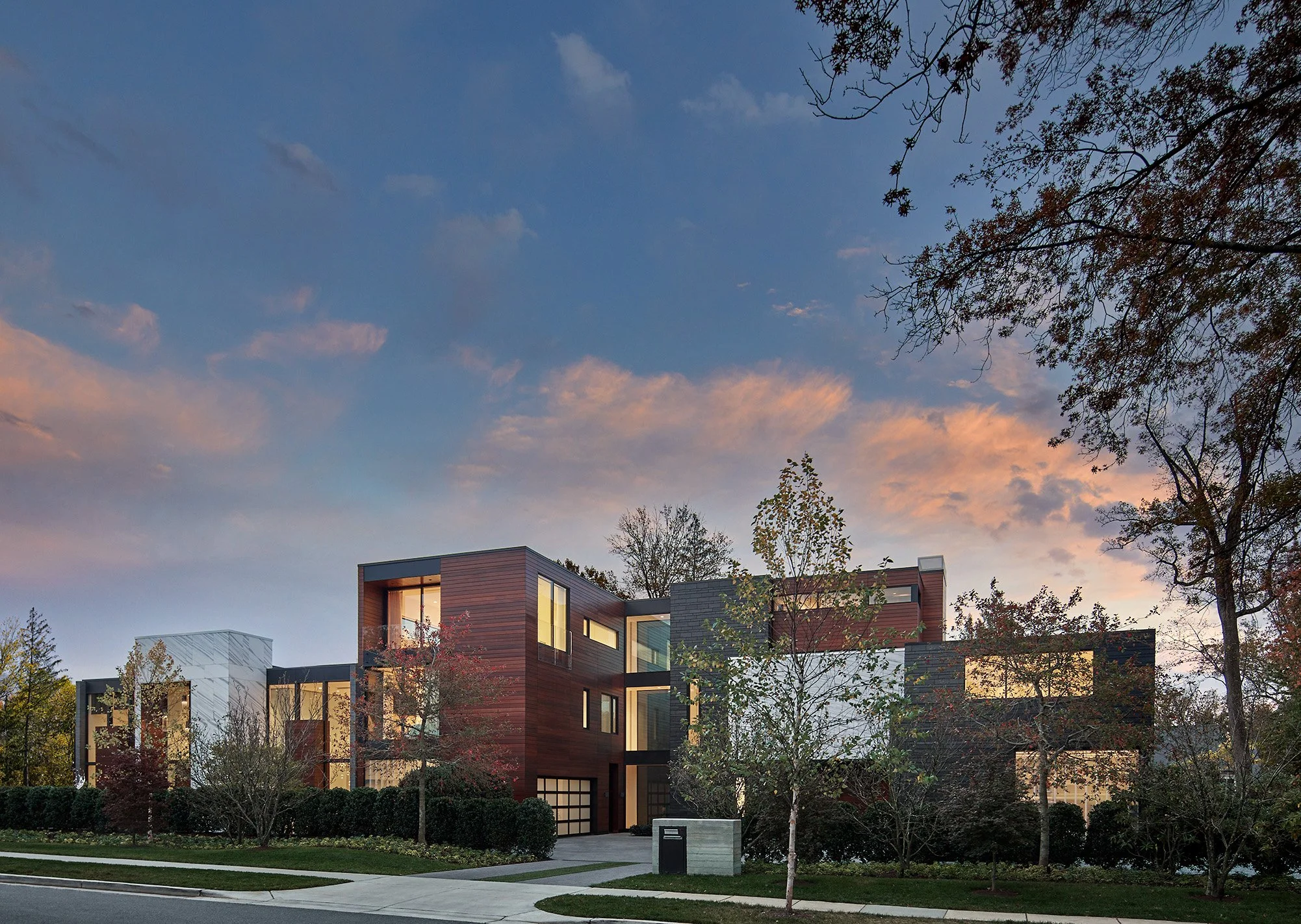 Modern multi-story residential buildings with large windows, surrounded by trees and a well-maintained lawn under a partly cloudy sky during sunset.