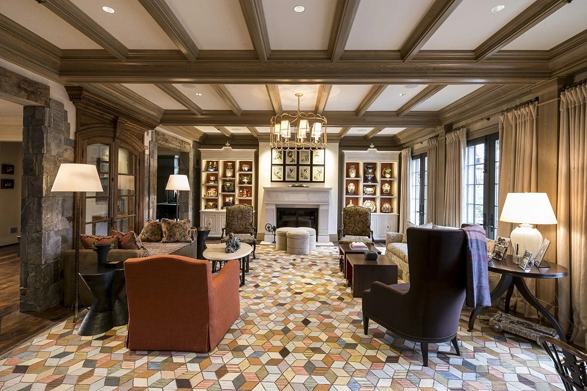 A spacious living room with a coffered ceiling, built-in shelves with decorative items, a fireplace, and multiple chairs and sofas arranged around a patterned rug.