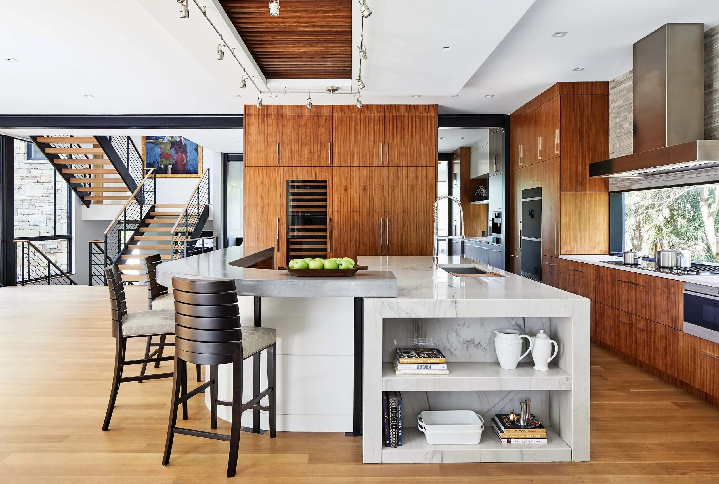 Modern open-concept kitchen with white marble island, wooden cabinetry, and staircase with black railing in the background.