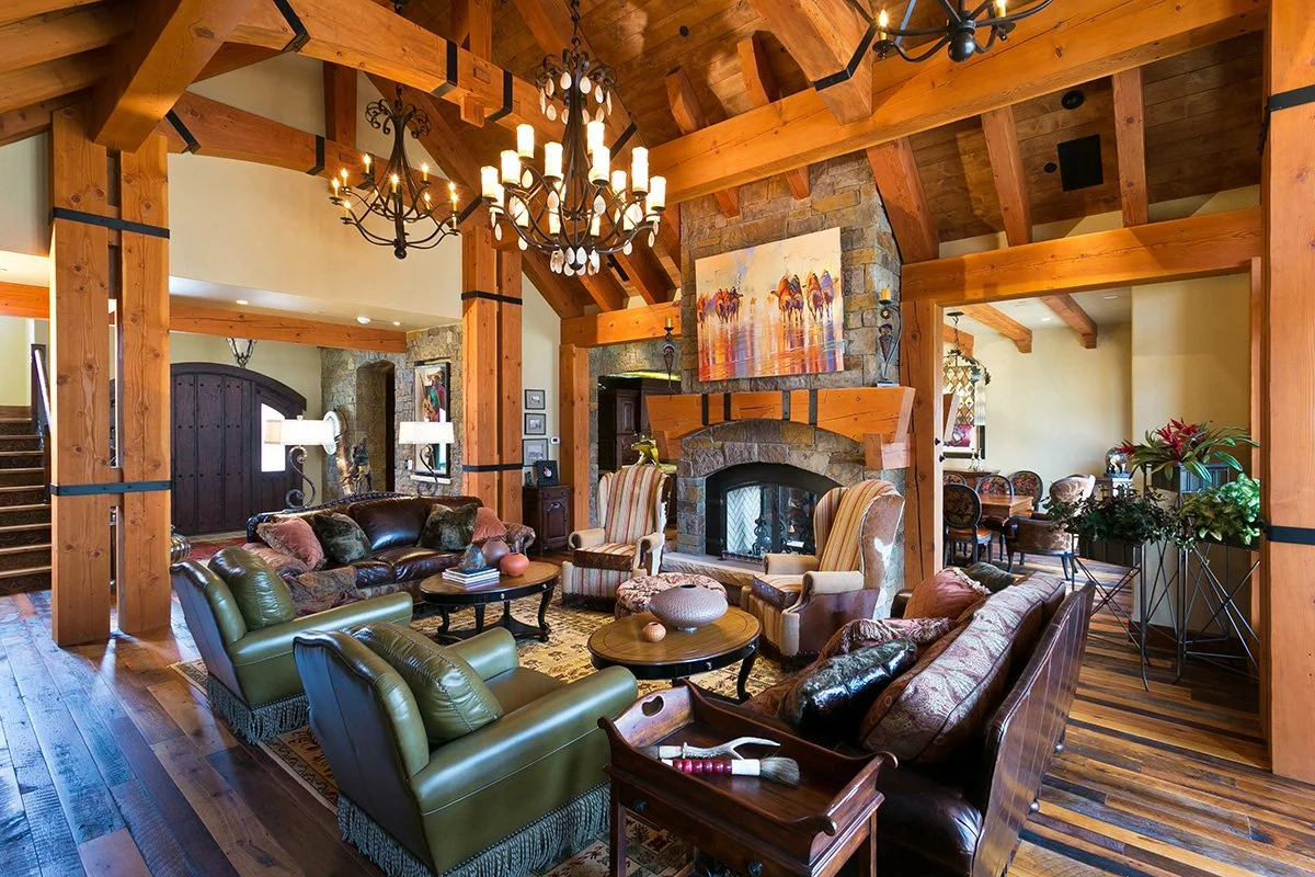 A rustic living room with wooden beams, a chandelier, and a stone fireplace. The room has various cushioned chairs and sofas arranged around a coffee table, with art on the walls and a dining area in the background.
